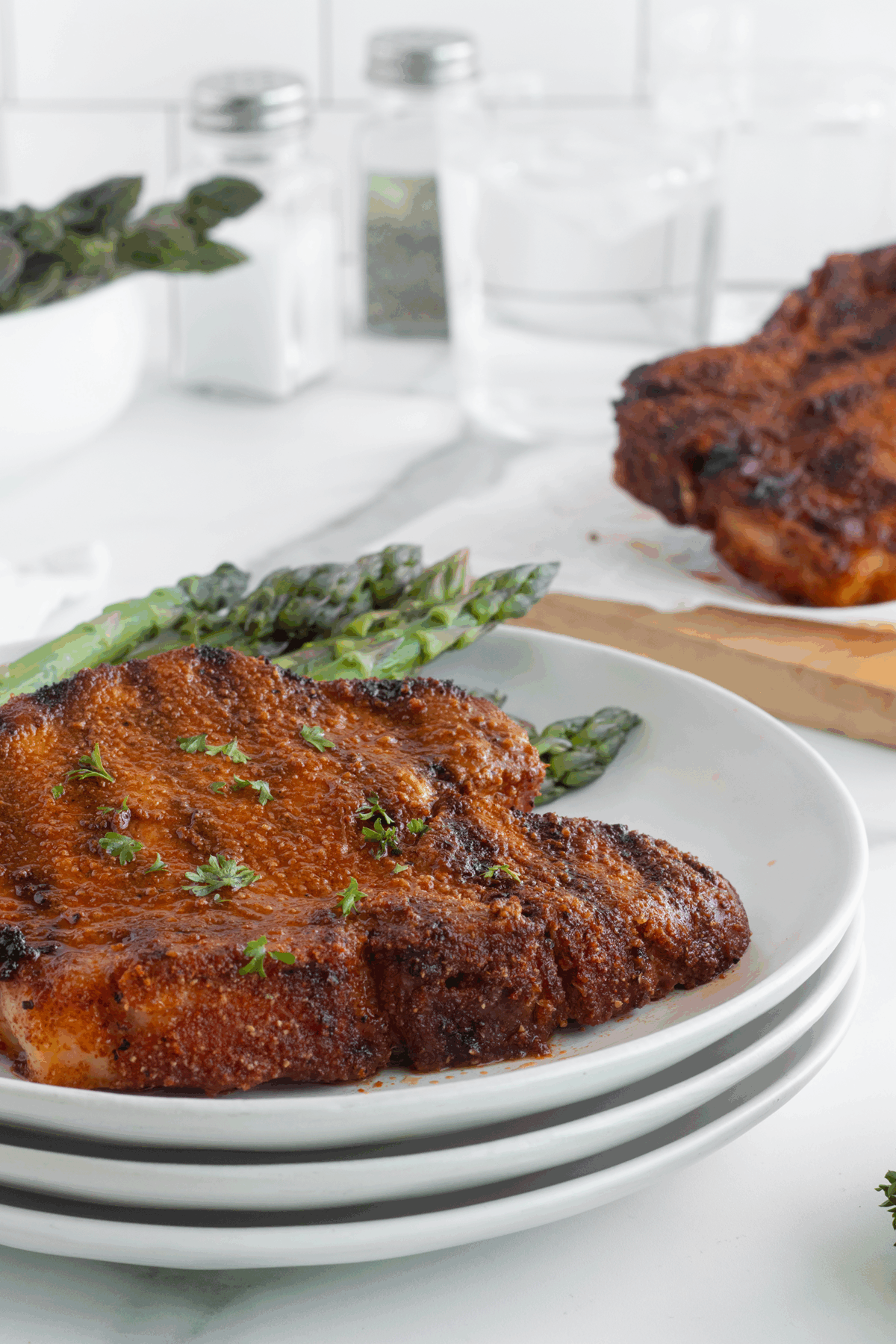 A seasoned, grilled pork chop garnished with herbs sits on a plate with asparagus, with another pork chop and spices visible in the background.