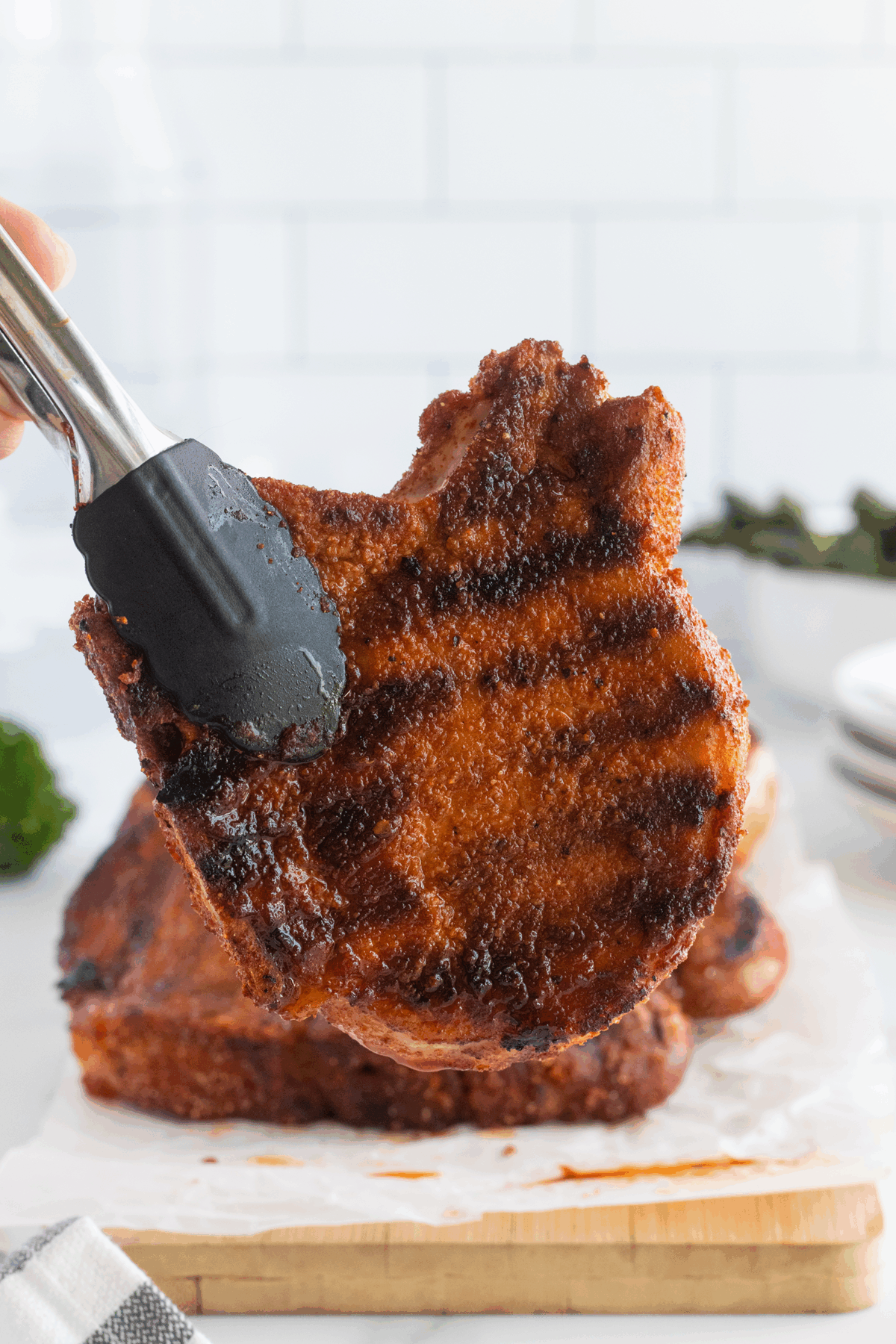 A pair of tongs holds up a grilled, seasoned pork chop in front of a stack of similar chops on a wooden cutting board, with a white tiled background.