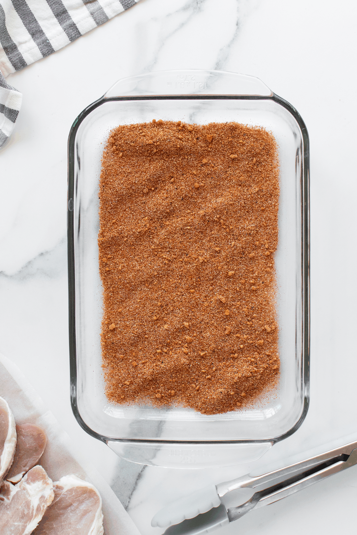 A glass baking dish filled with a reddish-brown dry spice rub sits on a white marble surface. Nearby are metal tongs, a striped cloth, and uncooked pork chops on a tray.