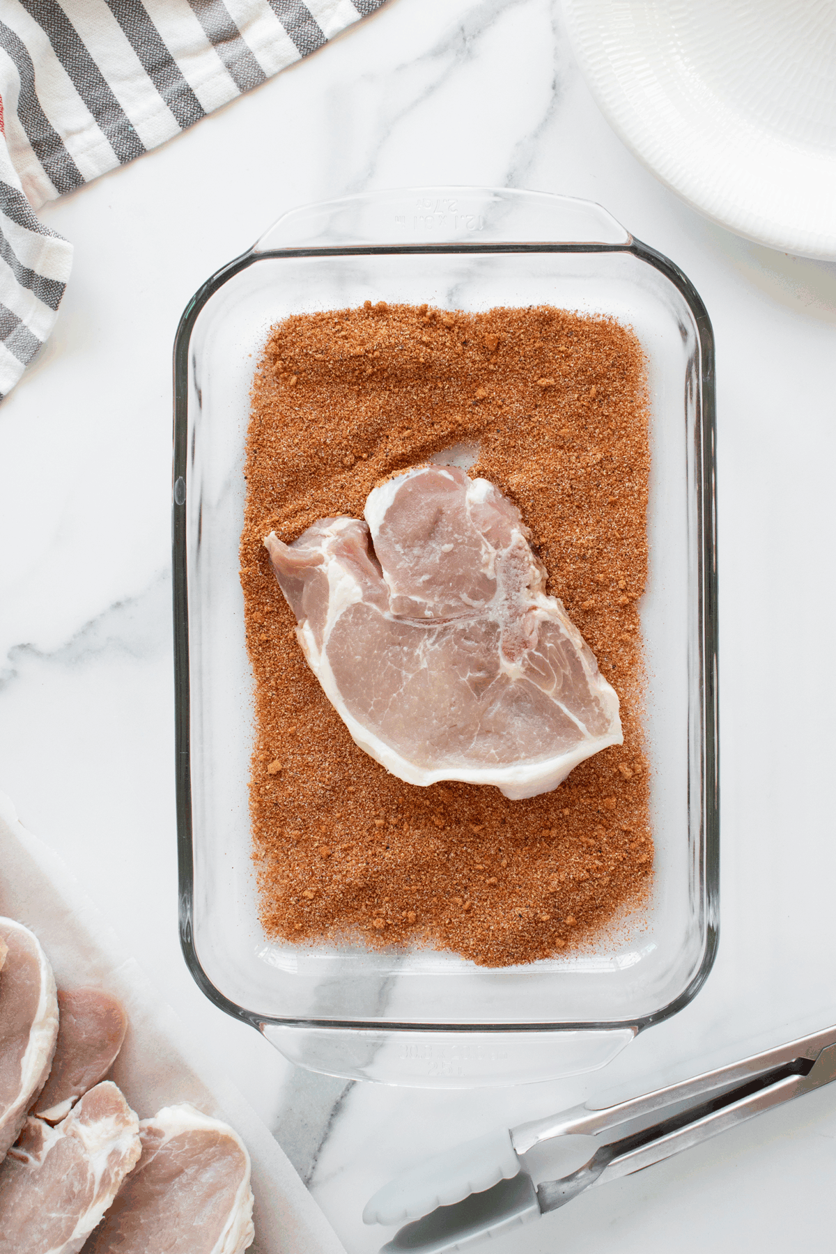 A raw pork chop sits on top of a layer of dry spice rub in a glass baking dish, with tongs, a plate, and more pork chops nearby on a white marble surface.