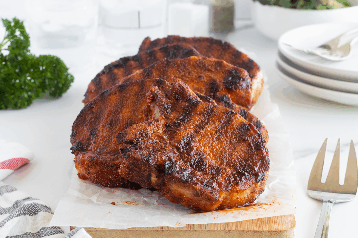 Three grilled pork chops with a dark, seasoned crust rest on parchment paper atop a wooden board, with plates, utensils, and parsley in the background.