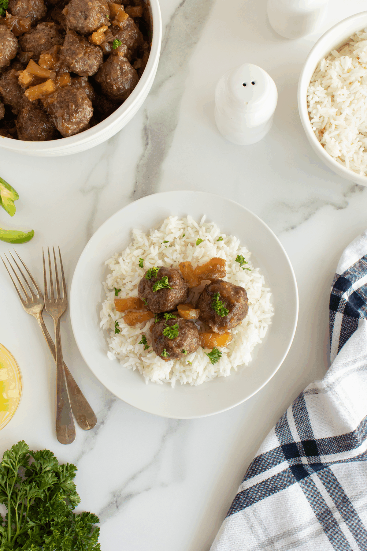 A plate of white rice topped with meatballs and sauce is on a table with a bowl of rice, a bowl of meatballs, parsley, two forks, salt and pepper shakers, and a blue plaid napkin.
