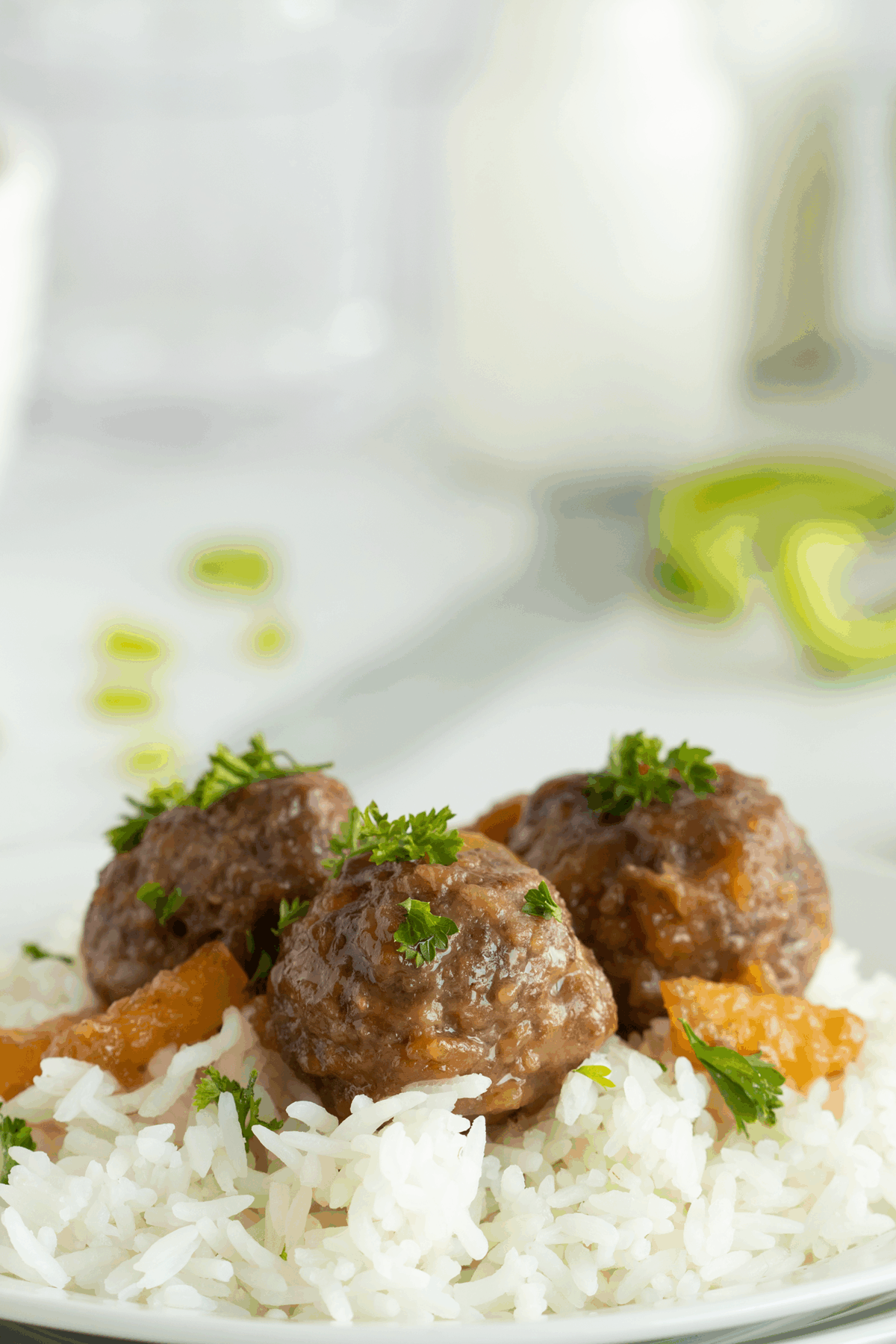 Three meatballs with sauce and herbs are served on a bed of white rice, garnished with parsley. The background is softly blurred, showing hints of green vegetables and a white bottle.