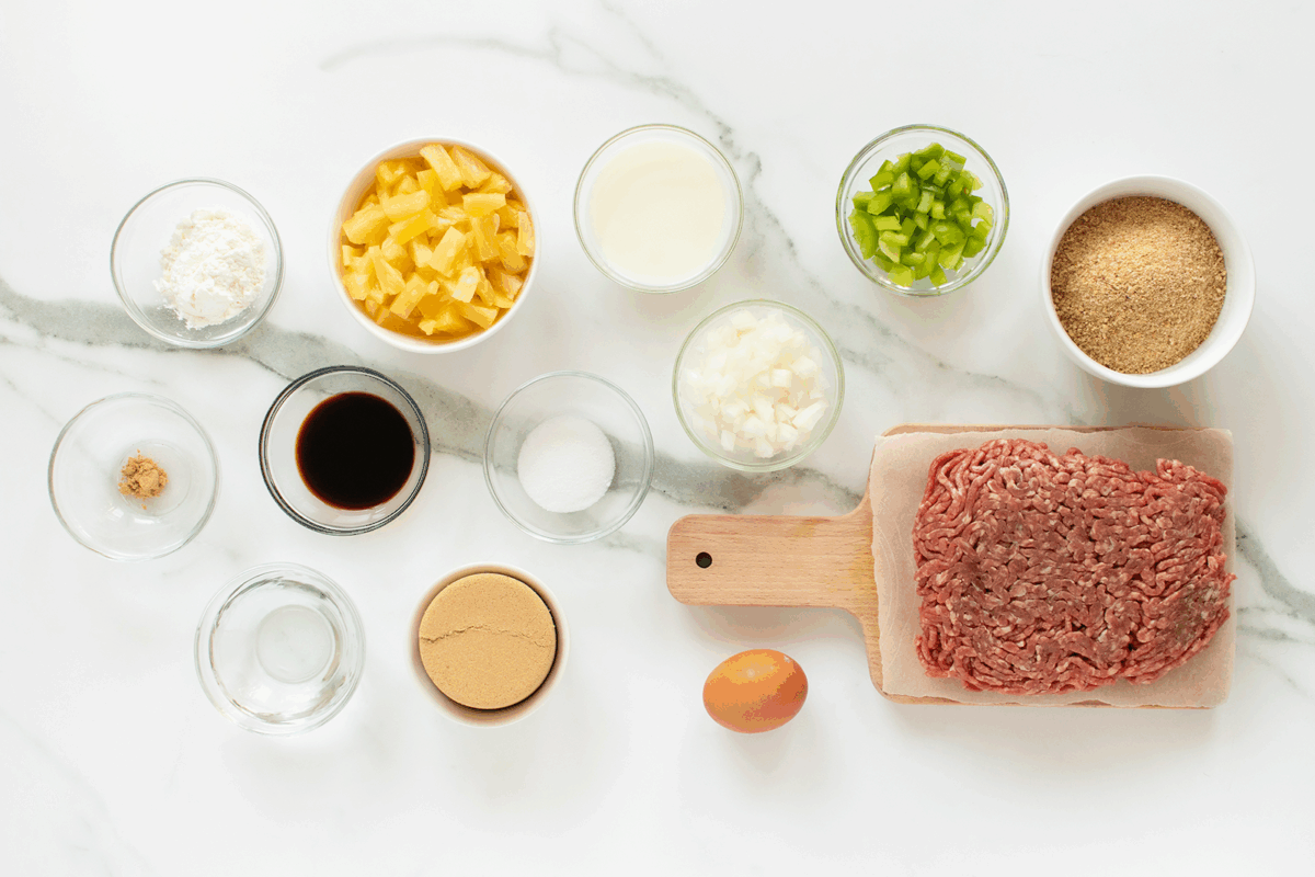 Various ingredients are arranged on a white marble surface, including ground beef, an egg, chopped onions and peppers, pineapple chunks, bread crumbs, brown sugar, soy sauce, flour, vinegar, milk, and seasonings in small bowls.