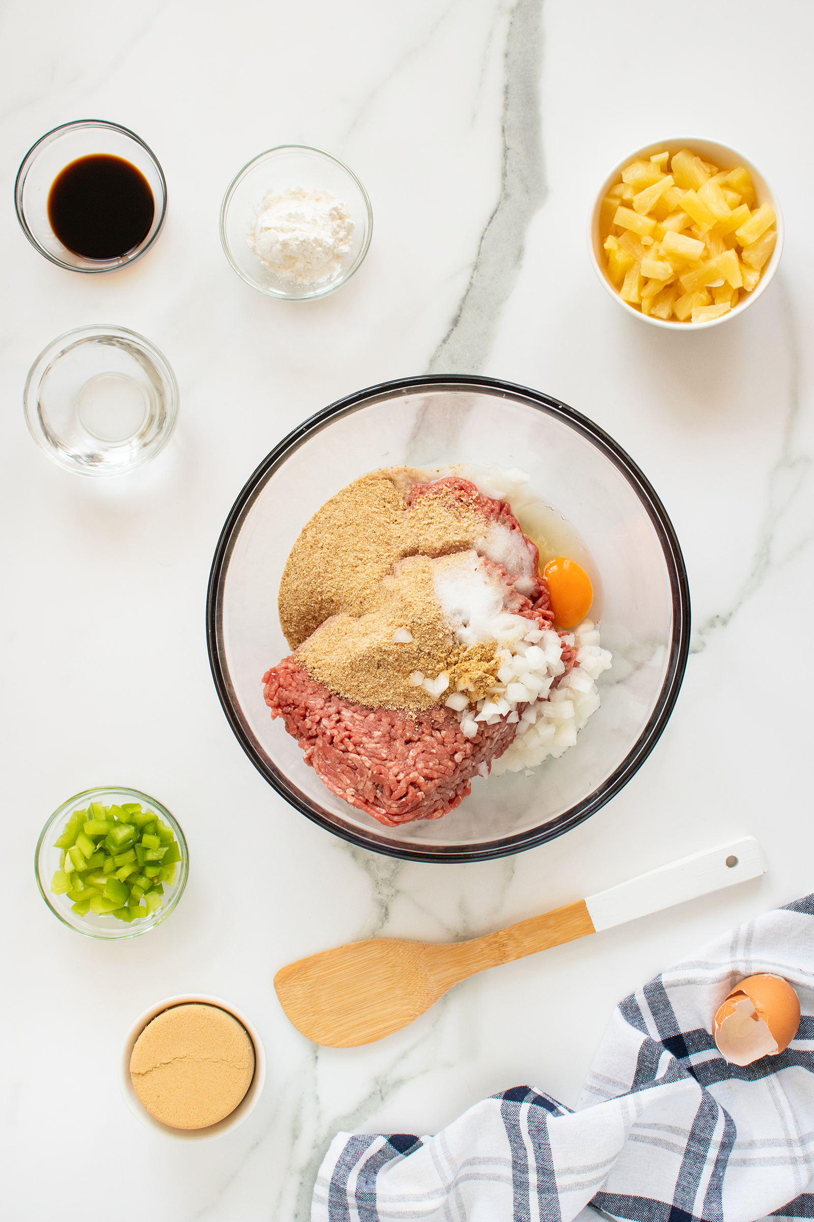 A glass mixing bowl with ground meat, breadcrumbs, chopped onion, an egg, and seasoning sits on a white marble surface. Surrounding it are small bowls with brown sugar, pineapple, green pepper, soy sauce, and other ingredients.