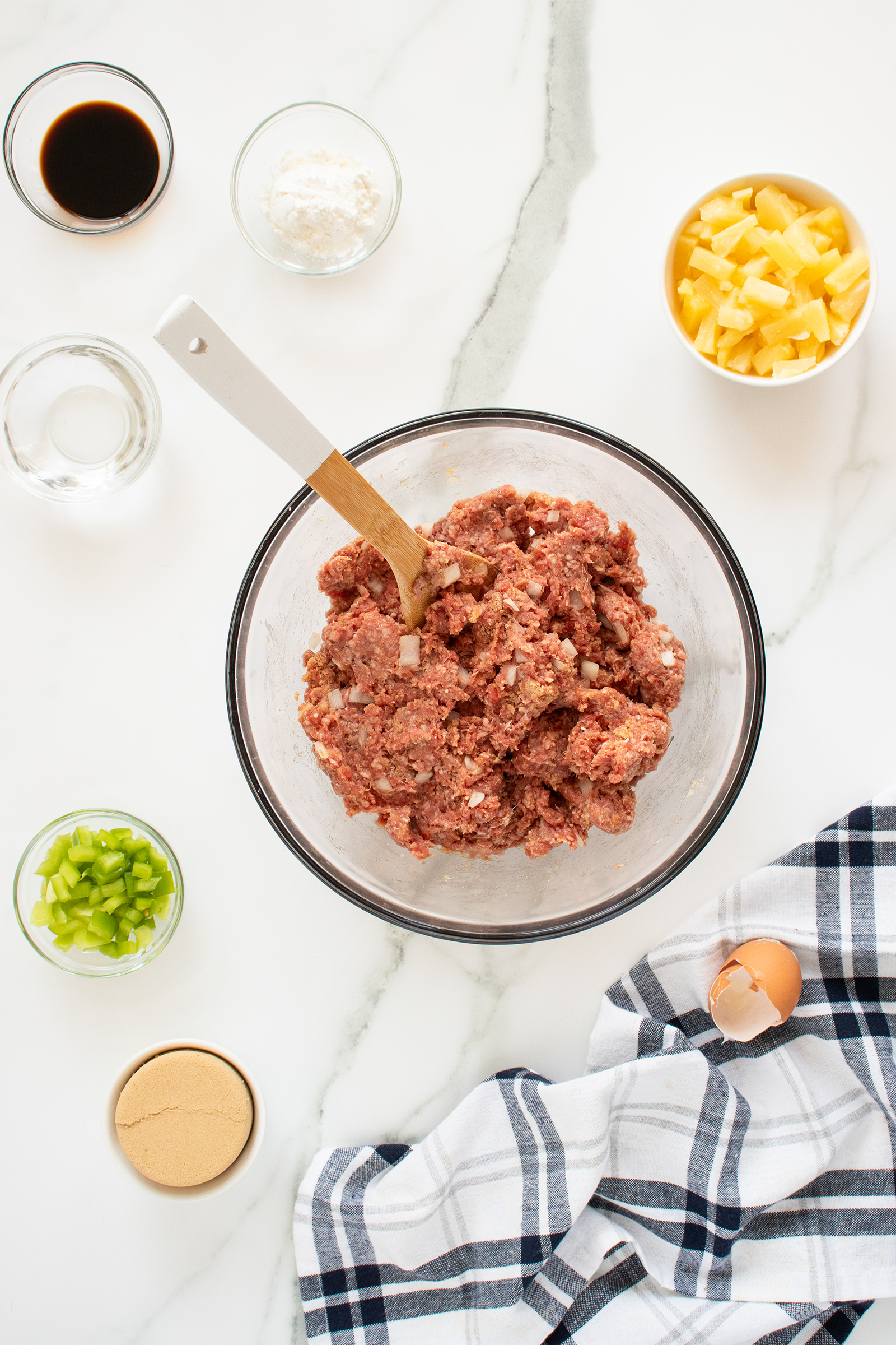 A glass bowl of ground meat being mixed with a spatula, surrounded by small bowls of brown sugar, chopped green pepper, pineapple chunks, cornstarch, soy sauce, water, and a cracked egg on a marble countertop with a plaid towel.