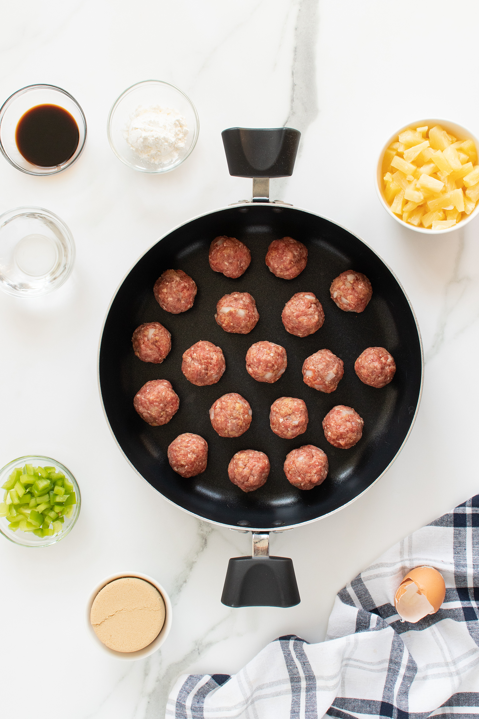 A top-down view of a skillet with uncooked meatballs, surrounded by small bowls of diced pineapple, chopped green pepper, brown sugar, cornstarch, soy sauce, and water, on a white marble surface with a striped towel and cracked egg.