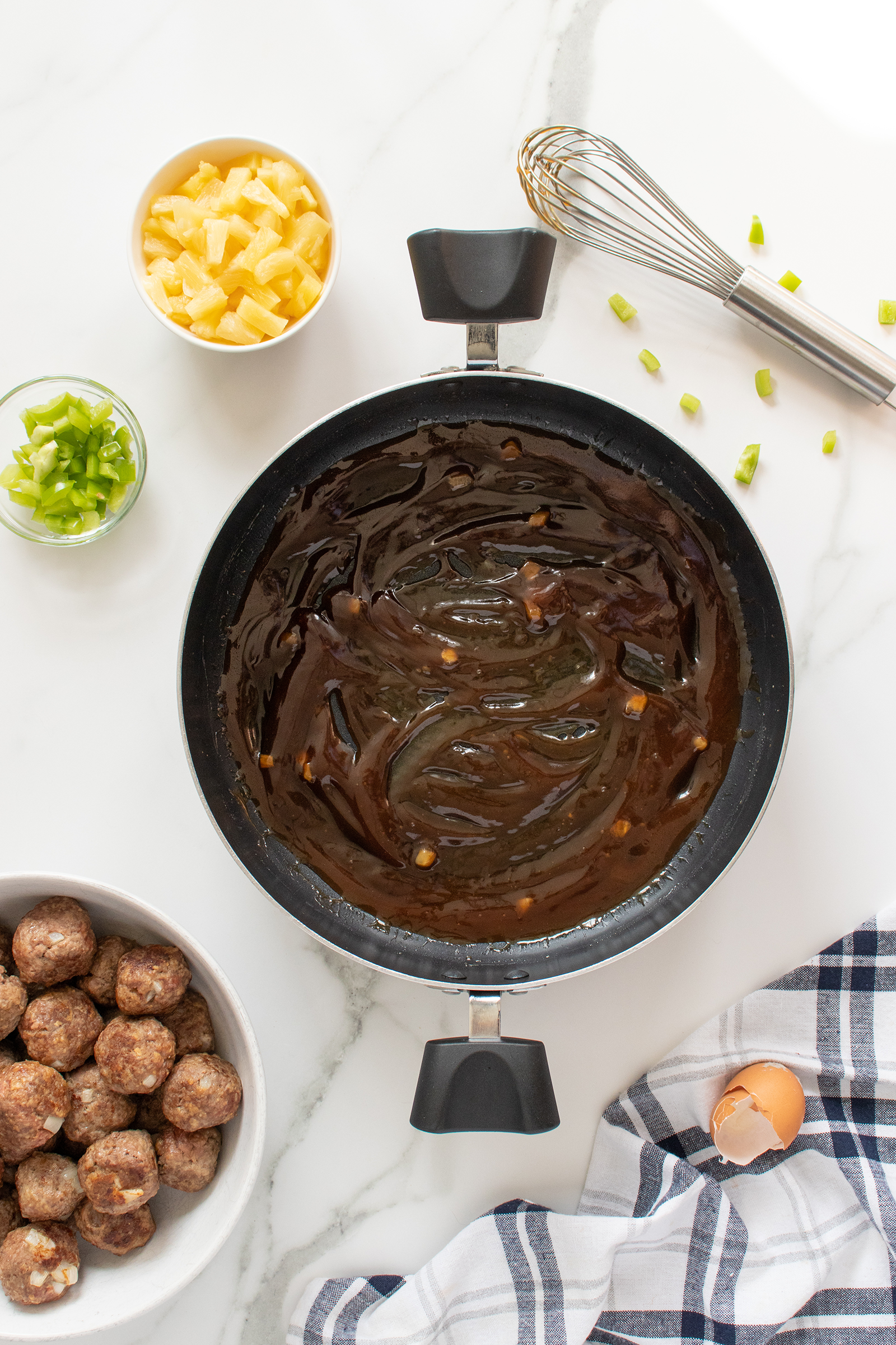 A saucepan with a dark sauce sits on a marble counter, surrounded by a bowl of meatballs, chopped green peppers, pineapple chunks, a whisk, a cracked egg, and a striped dish towel.