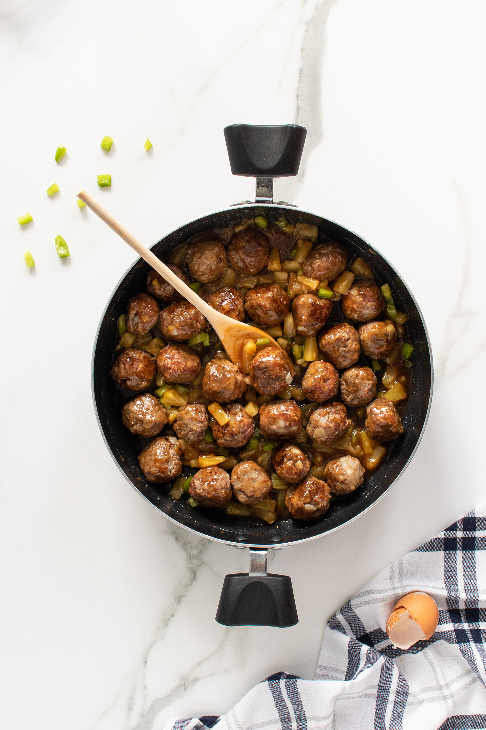 A black pot filled with glazed meatballs and chopped green peppers sits on a white marble surface. A wooden spoon rests in the pot. Scattered green onions, a striped towel, and a cracked eggshell are nearby.