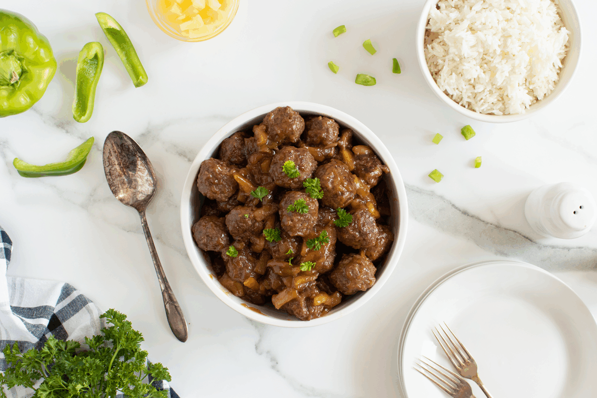 A bowl of meatballs in brown sauce with herbs sits on a marble surface, surrounded by a plate of white rice, green pepper slices, pineapple chunks, fresh parsley, a spoon, plates, and forks.