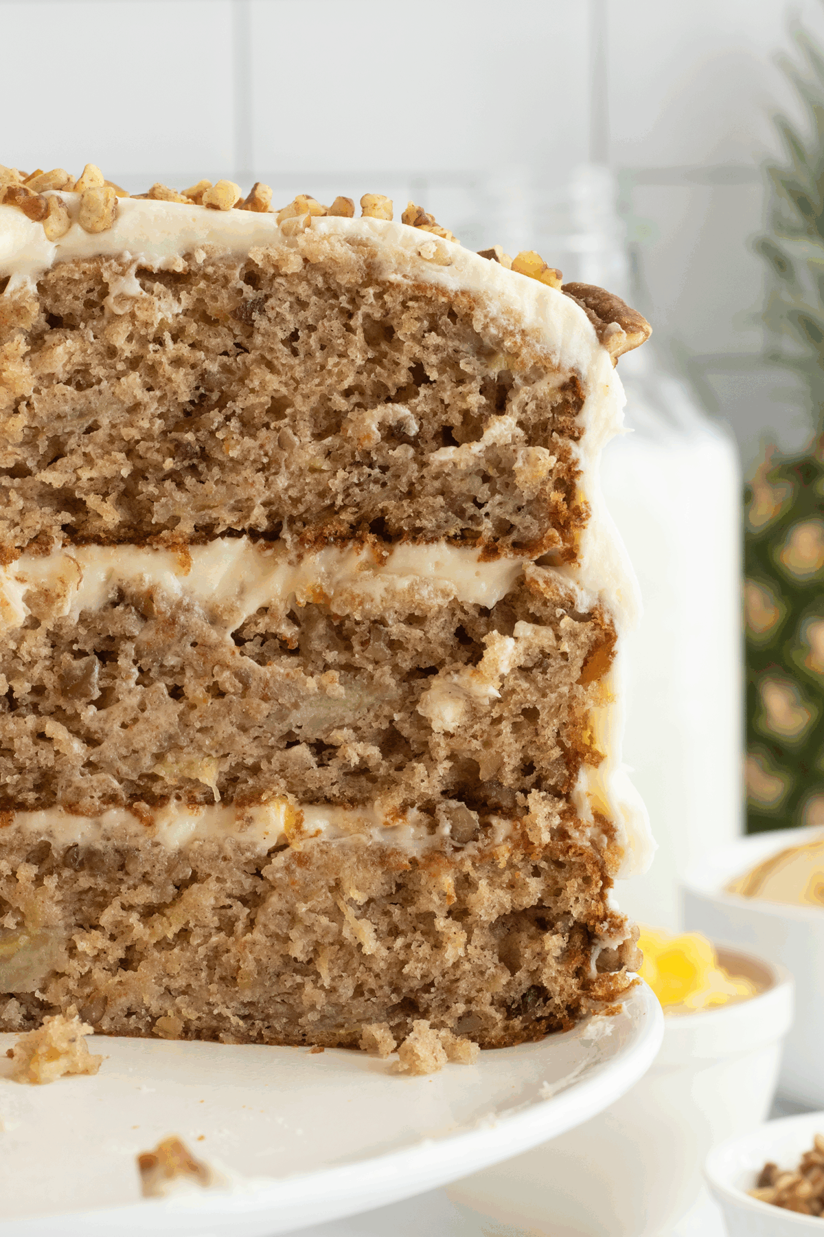 A close-up of a three-layer banana cake with cream cheese frosting and chopped nuts on top, displayed on a white cake stand. The cake appears moist and filled with banana pieces.