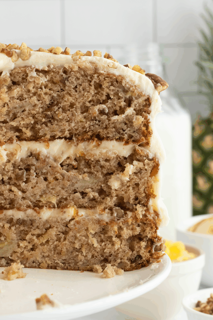 A close-up of a three-layer banana cake with cream cheese frosting and chopped nuts on top, displayed on a white cake stand. The cake appears moist and filled with banana pieces.