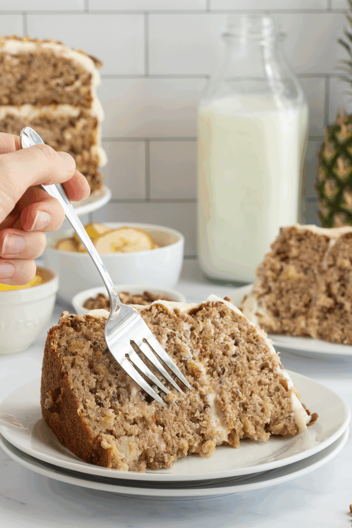 A hand holds a fork, about to take a bite of a slice of layered, frosted banana cake on a white plate. More cake, bananas, and a bottle of milk are visible in the background on a white countertop.