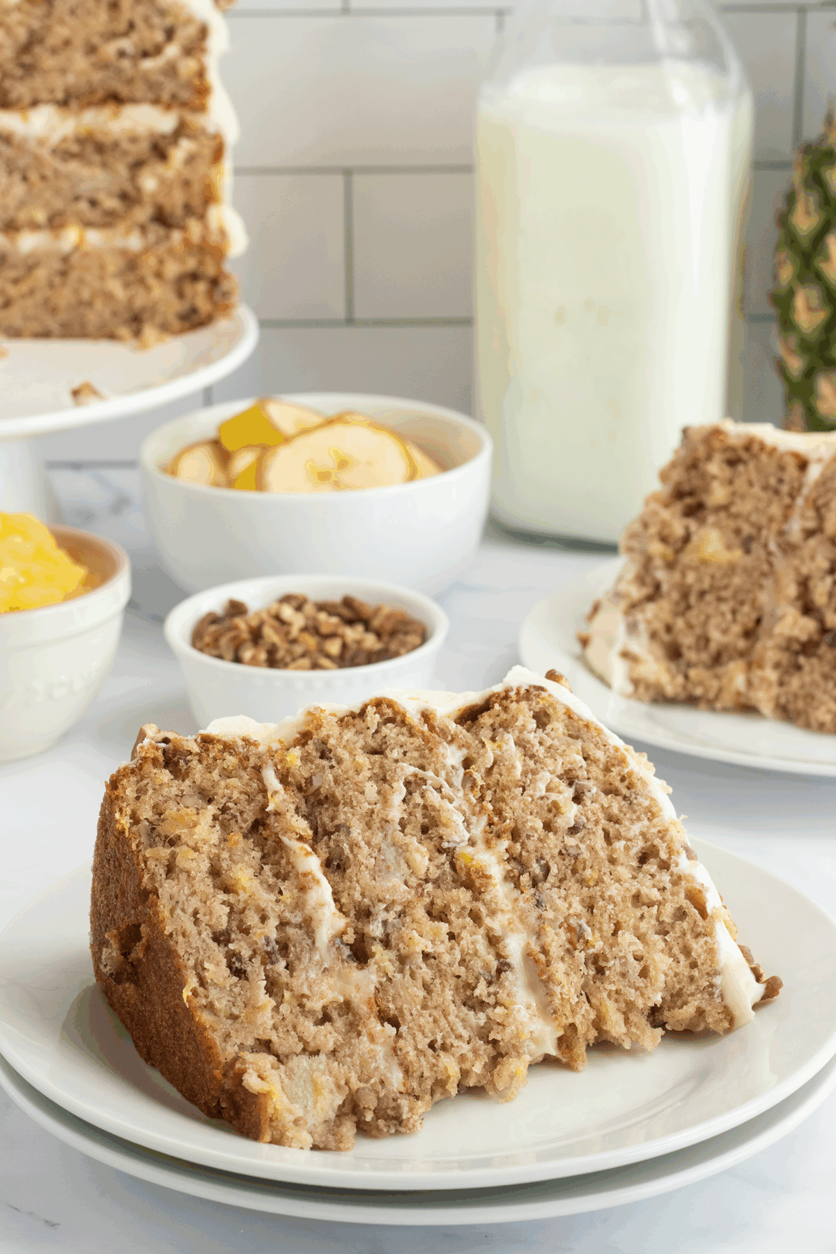 A slice of layered banana nut cake with frosting sits on a white plate, with another slice in the background. Nearby are bowls of bananas, pineapple, pecans, and a bottle of milk.