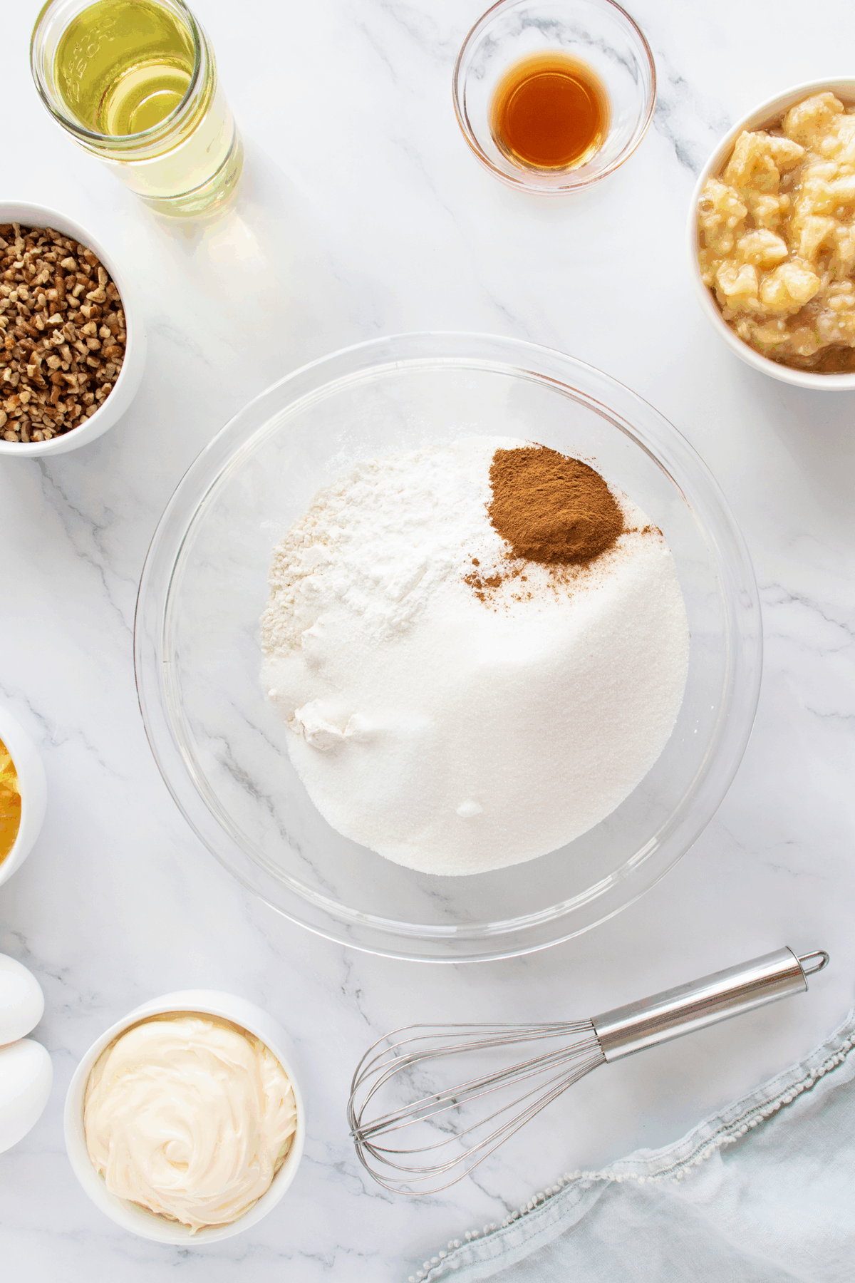 A bowl of flour, sugar, and cinnamon sits on a marble surface surrounded by bowls of mashed bananas, oil, nuts, vanilla, eggs, and mayonnaise, along with a whisk.