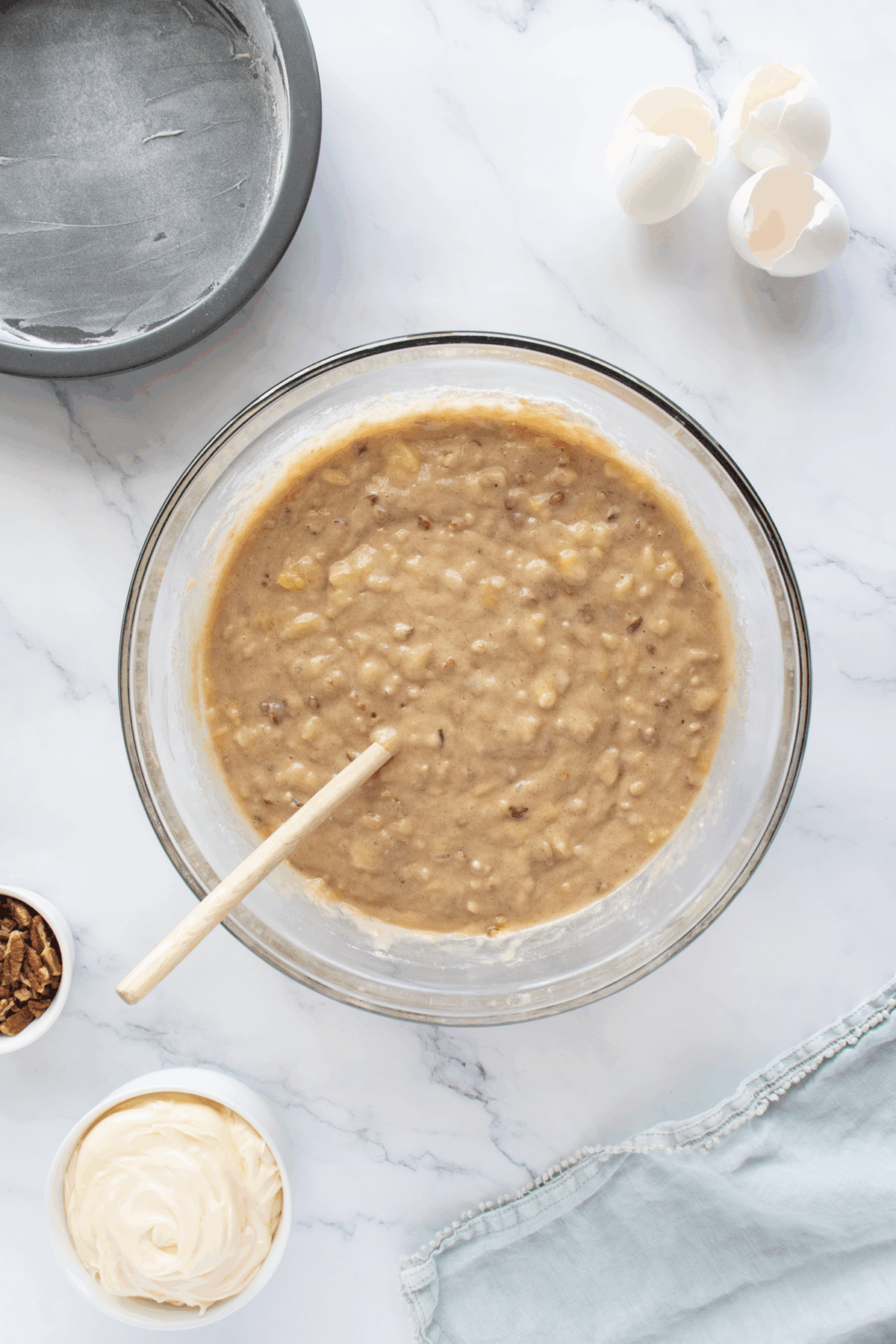 A glass bowl of banana bread batter with a wooden spoon sits on a marble counter. Nearby are cracked eggshells, a greased cake pan, a bowl of chopped nuts, a bowl of frosting, and a light blue cloth.