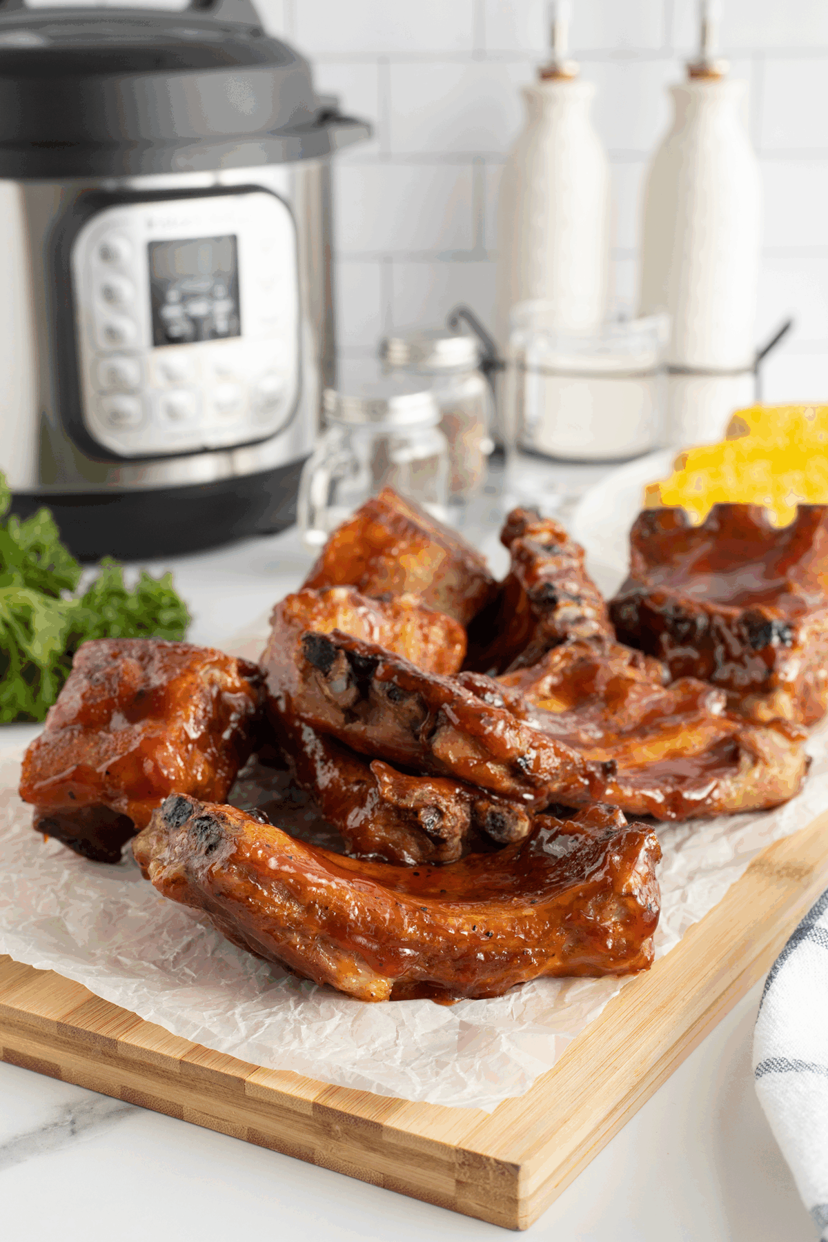 Barbecued ribs on parchment paper atop a wooden board, with an Instant Pot, seasoning jars, corn on the cob, and salt and pepper shakers visible in the background.