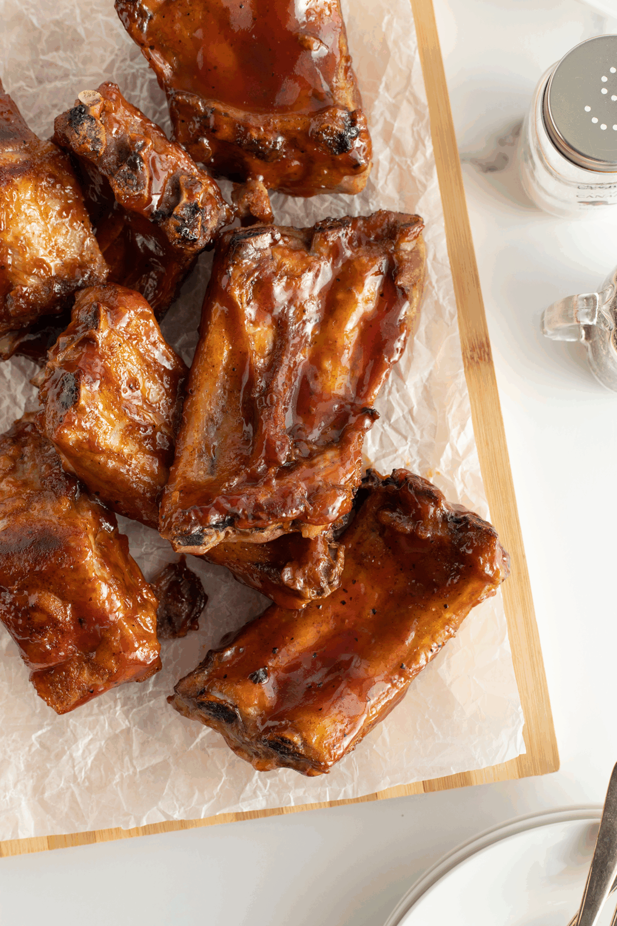 Barbecue pork ribs coated in a glossy sauce are arranged on parchment paper atop a wooden cutting board, with a salt shaker and dish visible in the background.