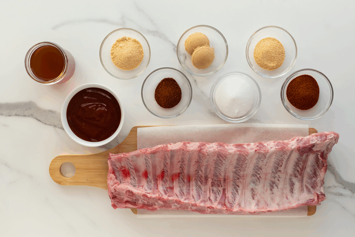 A rack of raw pork ribs on a wooden cutting board with small bowls of spices, sugar, brown sugar, barbecue sauce, and a jar of liquid on a white marble countertop.