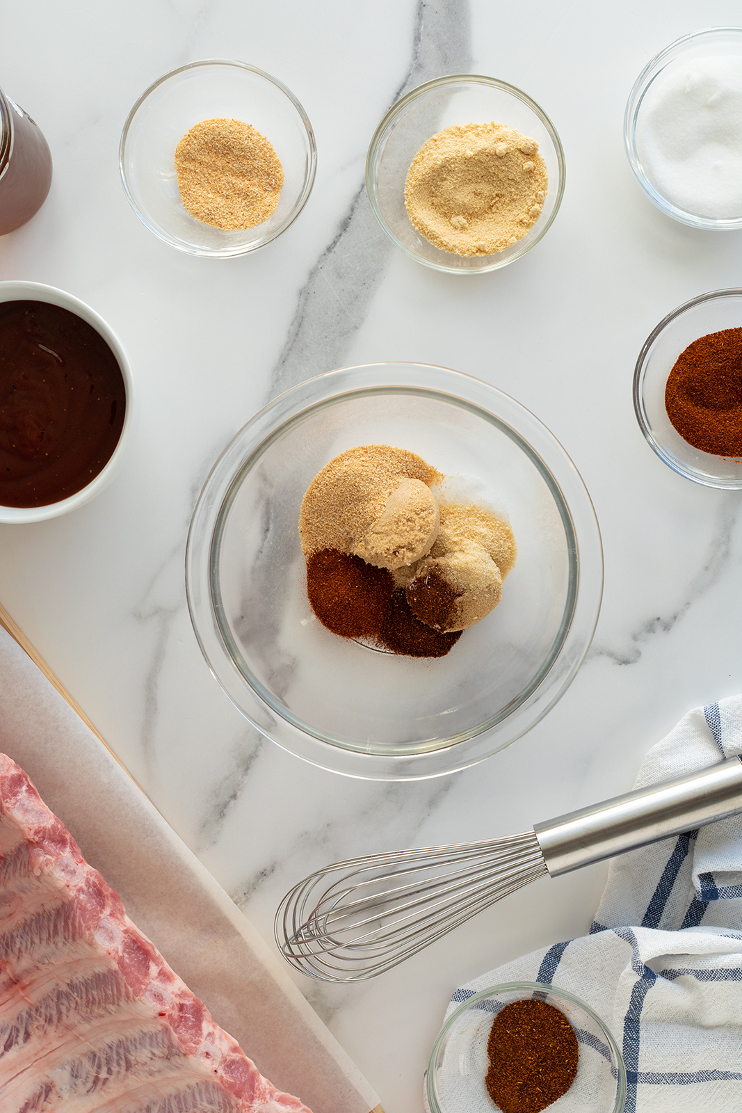 A glass bowl with brown spices and seasonings sits on a marble countertop, surrounded by small glass bowls of similar ingredients, a whisk, a striped towel, barbecue sauce, and a rack of raw ribs.