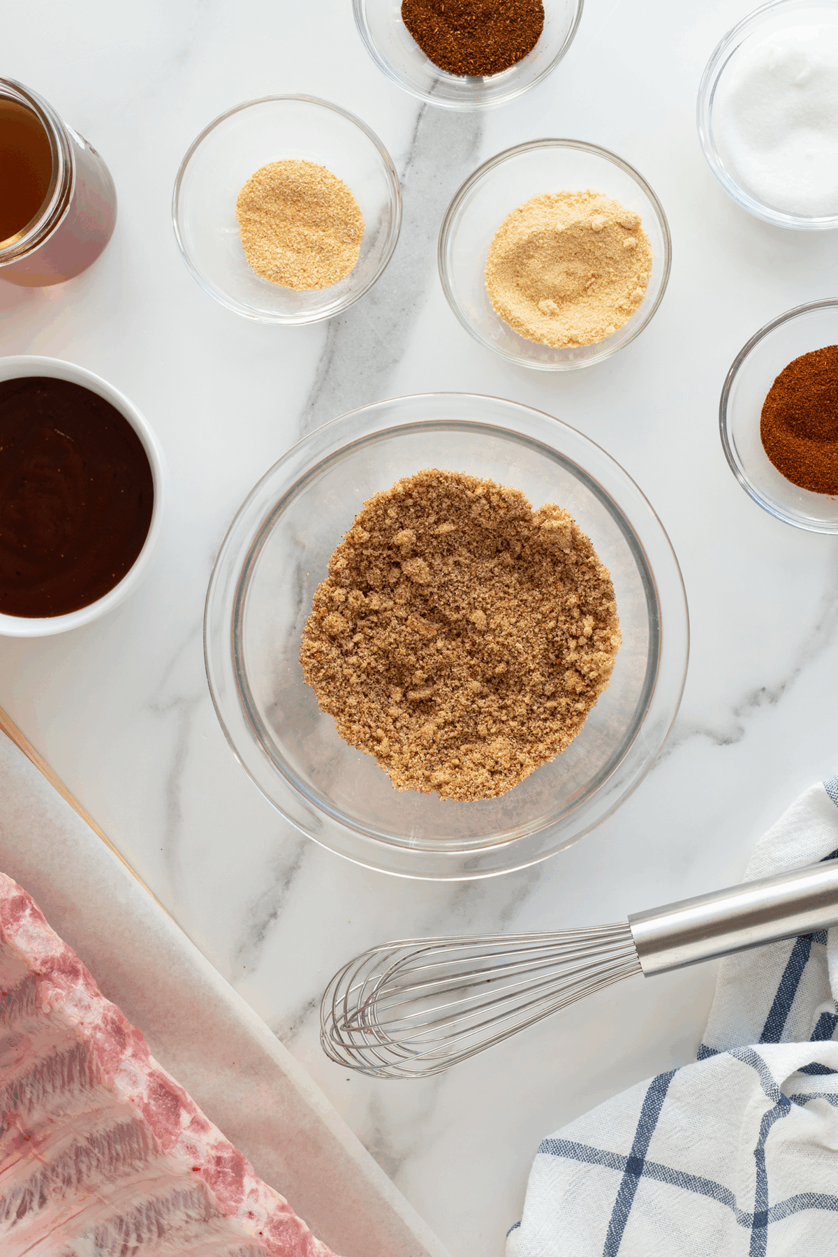 A glass bowl of brown sugar is surrounded by small bowls of spices, a sauce, and a jar of liquid on a white marble surface. Nearby are uncooked ribs on parchment paper, a metal whisk, and a blue-striped towel.