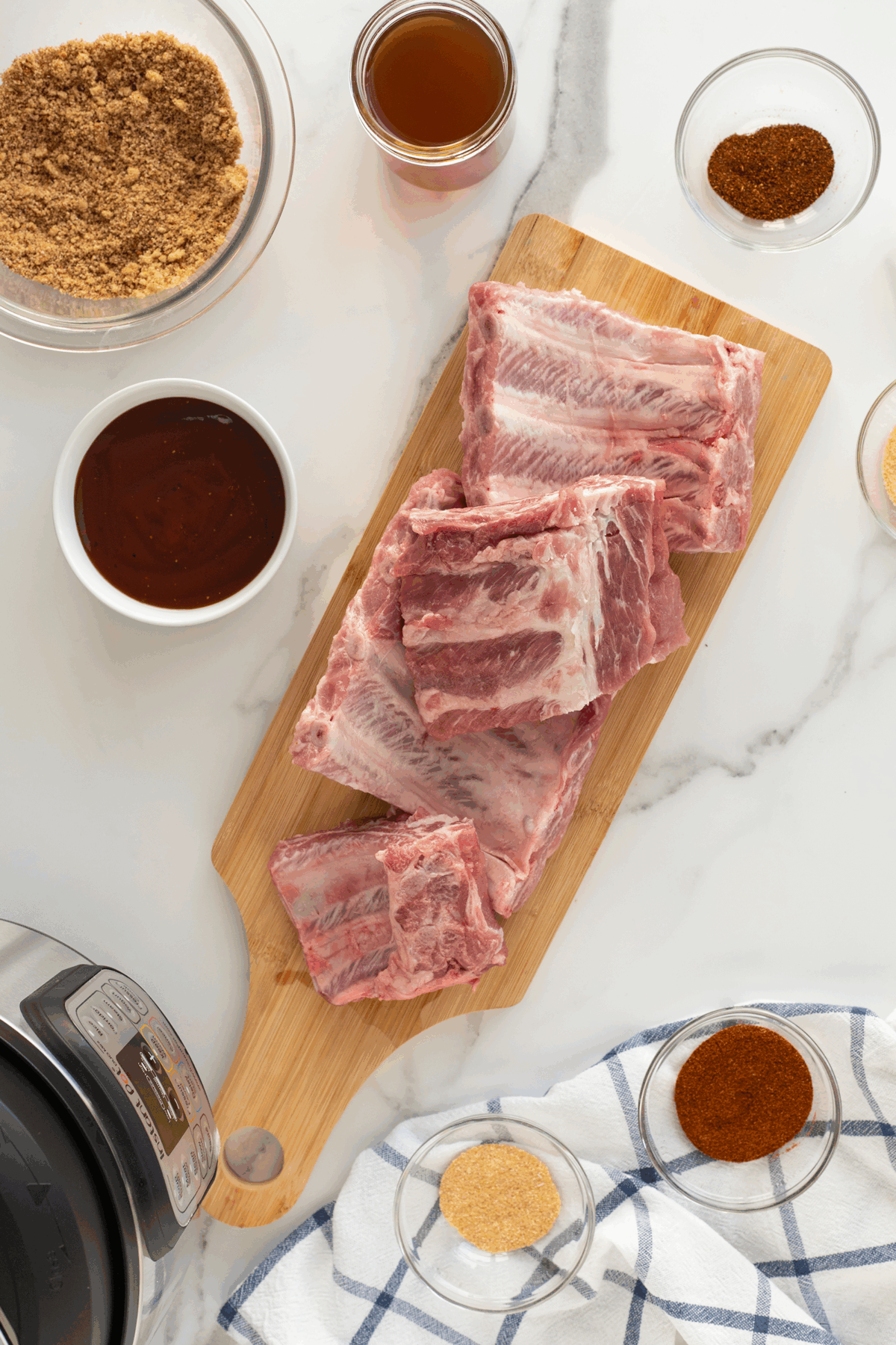 Raw pork ribs are stacked on a wooden cutting board, surrounded by bowls of brown sugar, spices, barbecue sauce, and broth on a white marble surface. An Instant Pot and a striped towel are also visible.