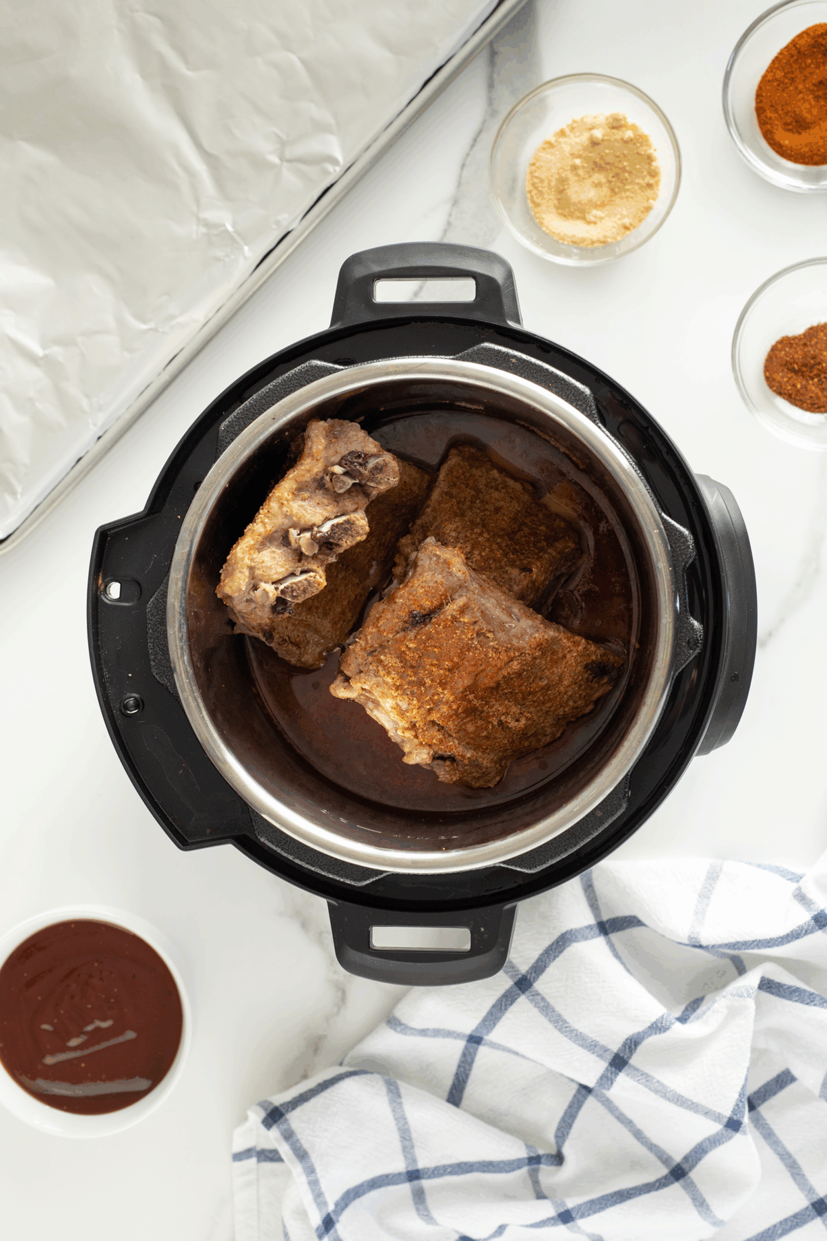 Overhead view of seasoned ribs cooking in an Instant Pot, surrounded by small bowls of spices, a bowl of barbecue sauce, a glass baking dish with foil, and a blue-striped kitchen towel on a white surface.