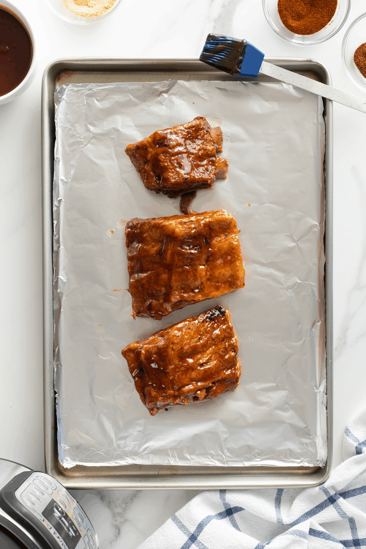 Three pieces of cooked ribs coated with sauce are arranged on a foil-lined baking sheet. A basting brush and bowls of spices and sauce are nearby on a white marble surface.