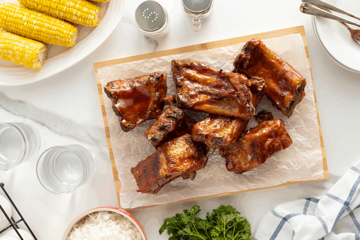 Barbecue ribs on a wooden board lined with parchment paper, surrounded by plates of corn on the cob, a bowl of rice, fresh parsley, salt and pepper shakers, glasses, and utensils on a white table.