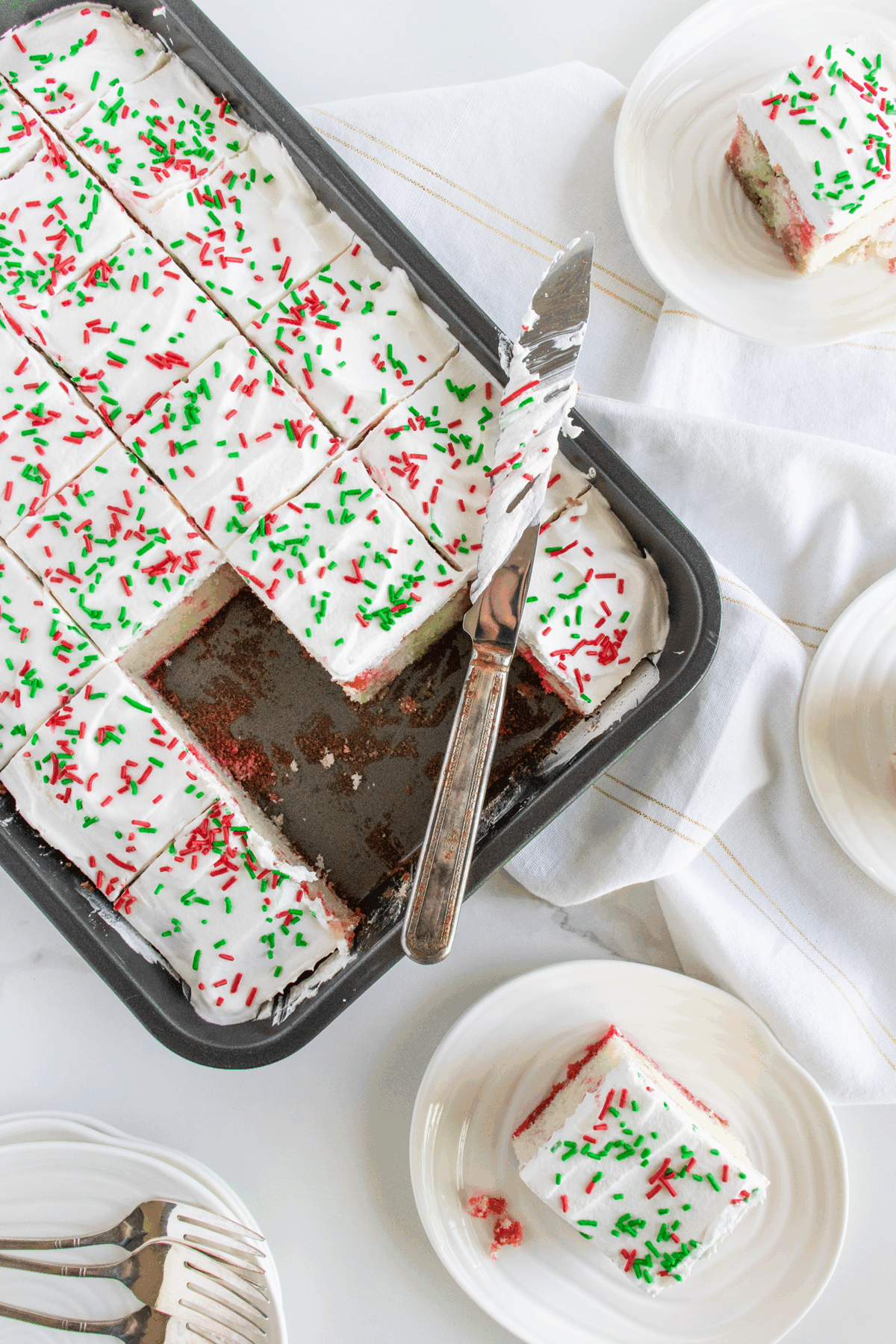 A baking pan of frosted sheet cake with red and green sprinkles, cut into squares. Several pieces are served on white plates nearby, with a serving knife resting in the pan.