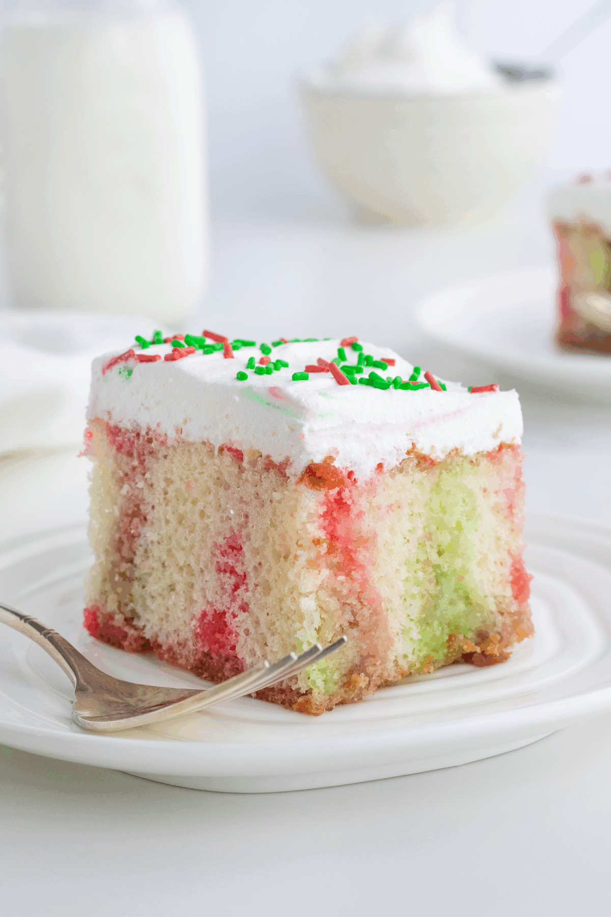 A slice of festive poke cake with white frosting, red and green sprinkles, and visible red and green swirls, served on a white plate with a fork. A blurred slice and dishes are in the background.