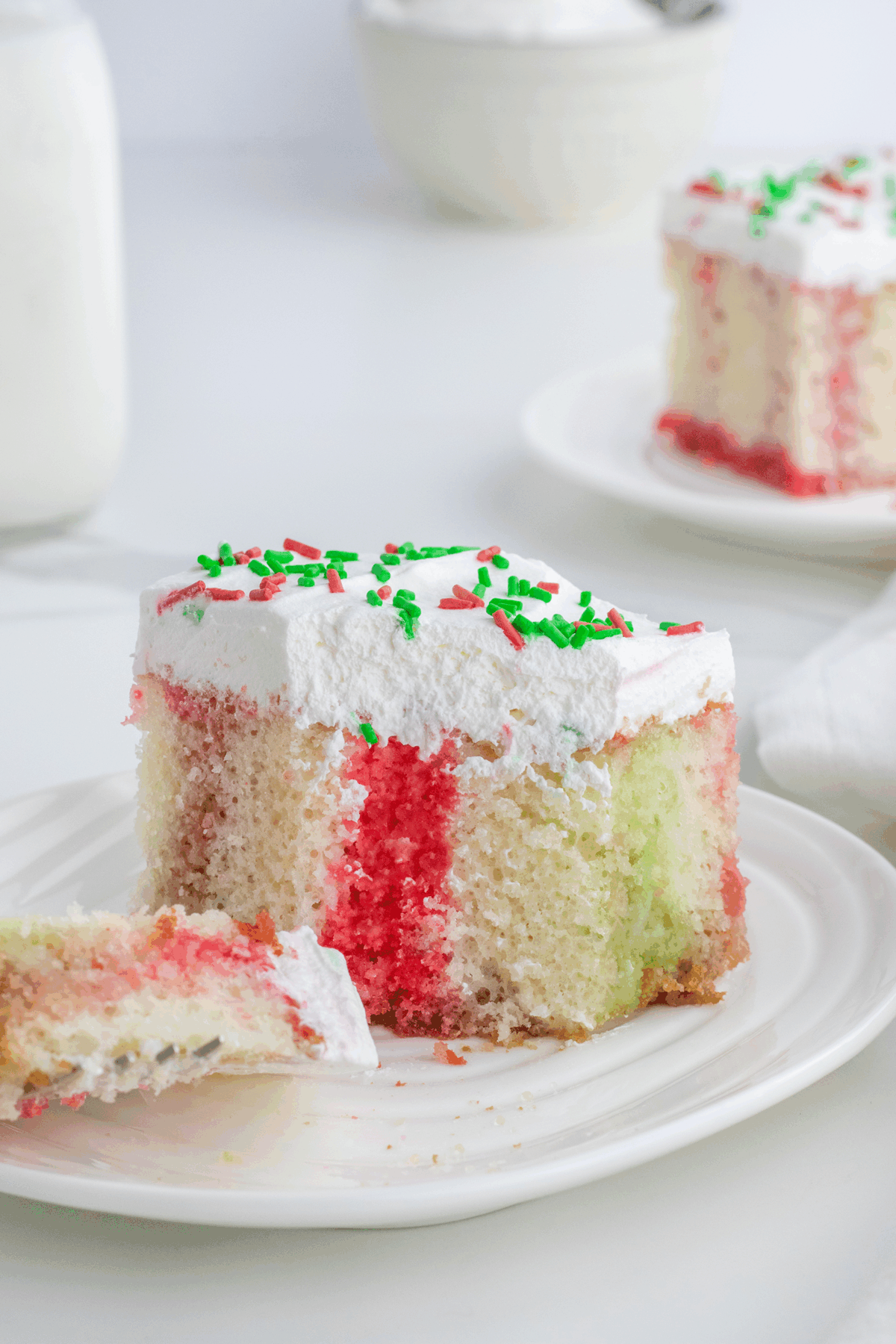 A slice of white cake with red and green swirled colors, topped with white frosting and red and green sprinkles, sits on a white plate with a forkful taken out. Another slice is in the background.