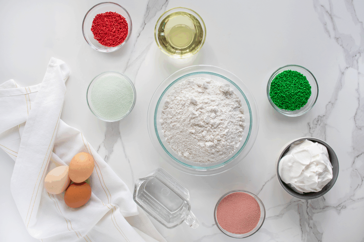 Baking ingredients on a white marble surface: flour in a bowl, eggs, oil, water, whipped cream, red and green sprinkles, pink powder, and a bowl of white powder. A white cloth napkin lies beside the eggs.