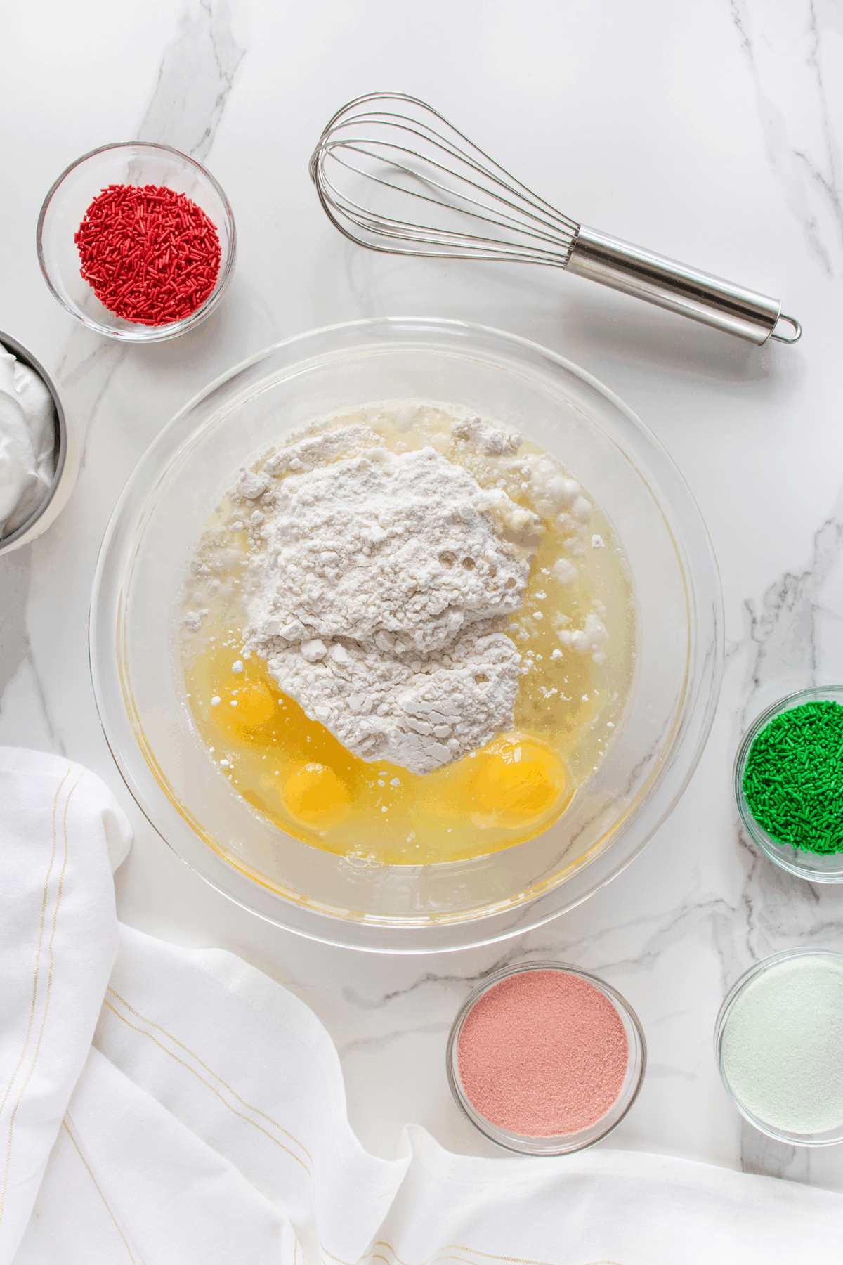 A glass bowl with eggs, flour, and wet ingredients sits on a marble countertop, surrounded by bowls of red and green sprinkles, pink sugar, and a metal whisk. A white cloth is nearby.