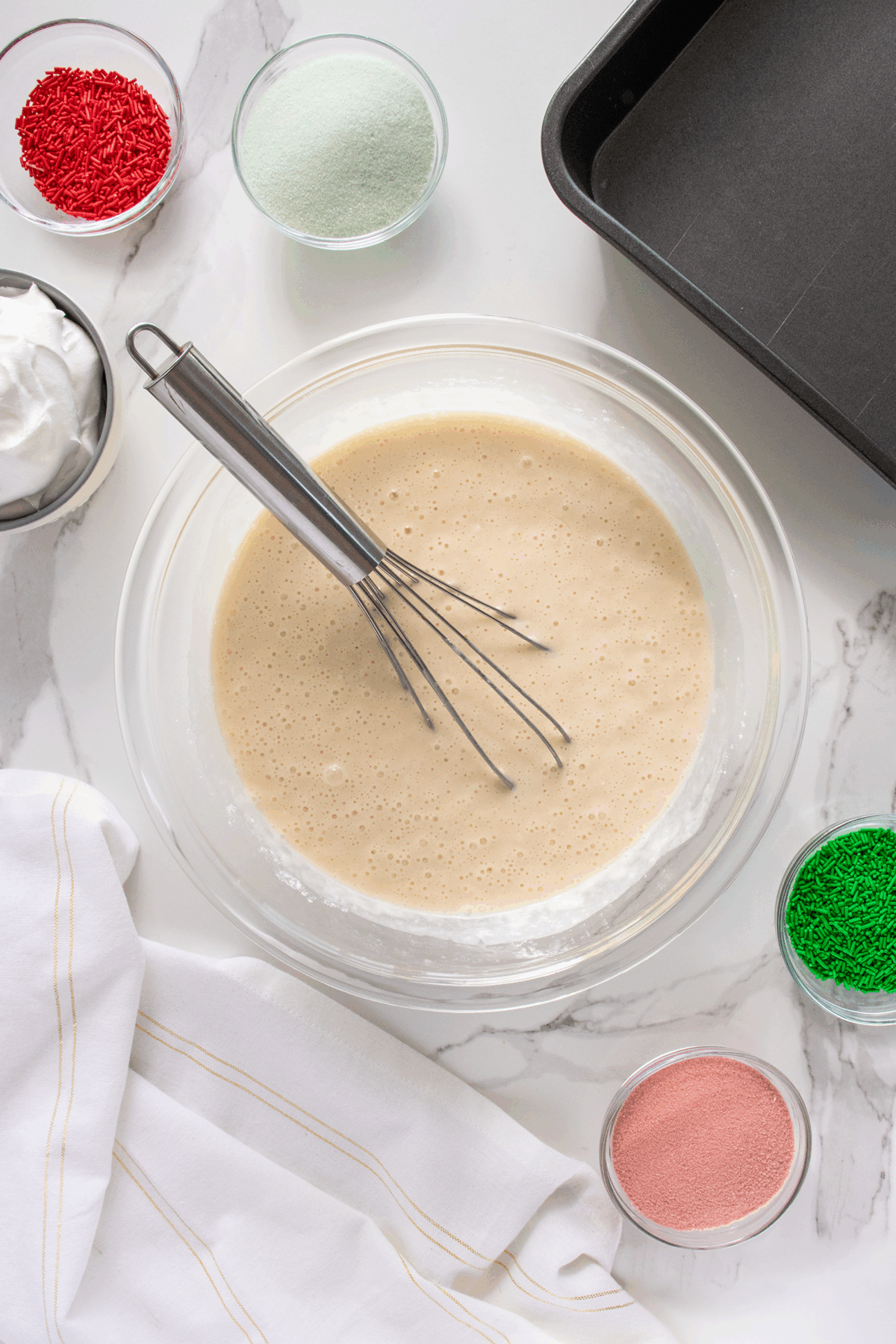 A glass bowl of cake batter with a whisk inside sits on a marble counter, surrounded by bowls of red and green sprinkles, white powder, pink powder, a baking pan, and a white kitchen towel.