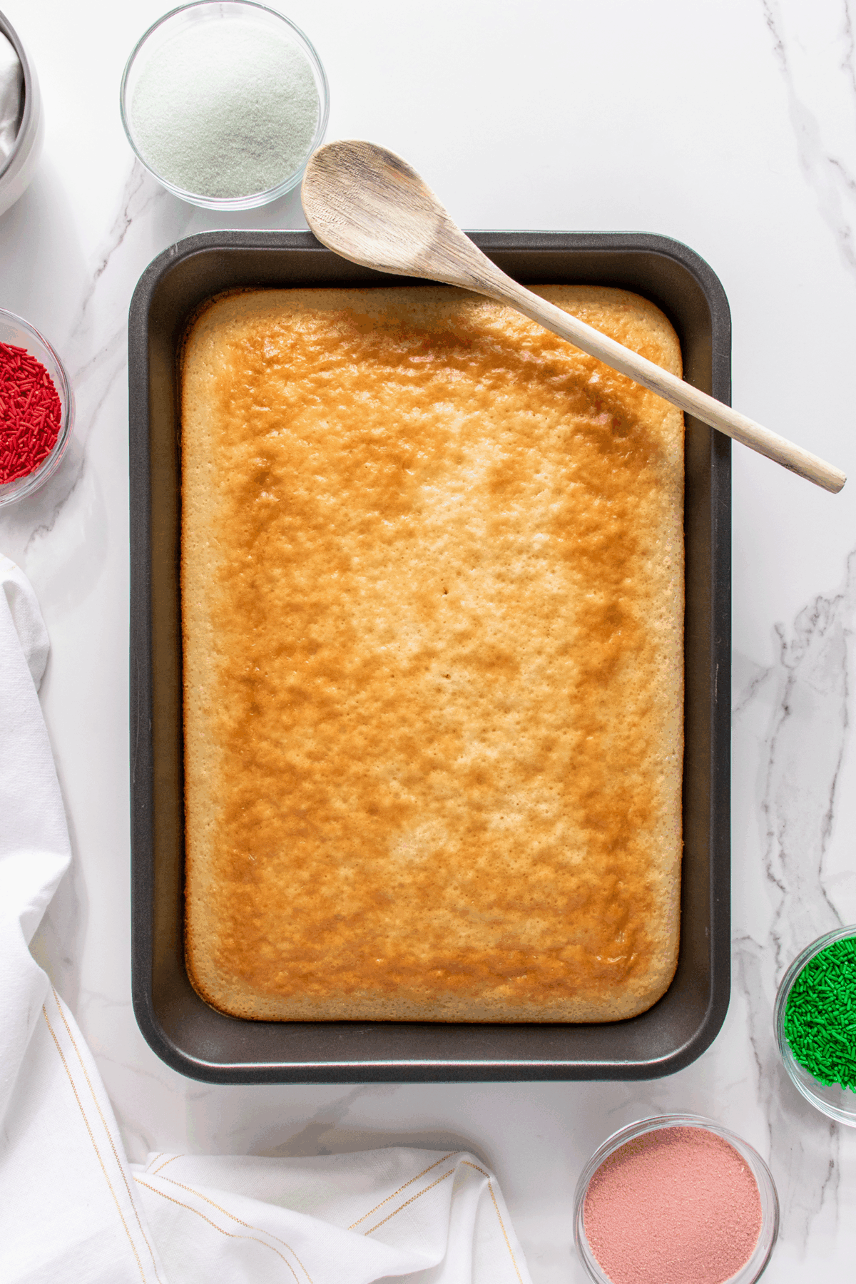 A golden-brown rectangular cake in a baking pan with a wooden spoon on top, surrounded by bowls of red, green, and pink sprinkles, and a white towel on a marble surface.