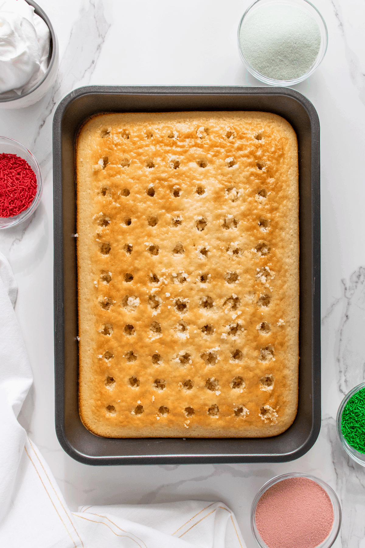 A rectangular cake with holes poked in it sits in a baking pan, surrounded by bowls of red and green sprinkles, white frosting, and green and pink powders on a marble surface.