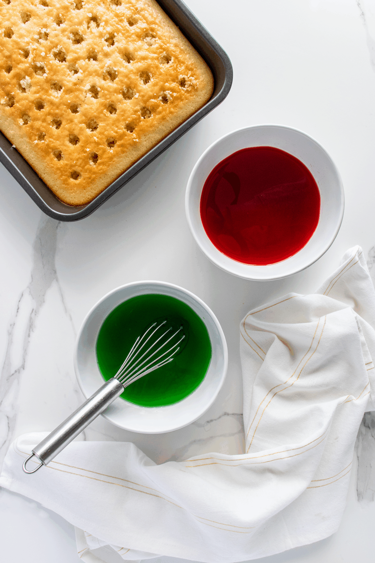 A baked rectangular cake with holes on top sits in a pan next to two bowls, one with red liquid and one with green liquid. A whisk rests in the green bowl, and a white cloth is nearby on a marble surface.