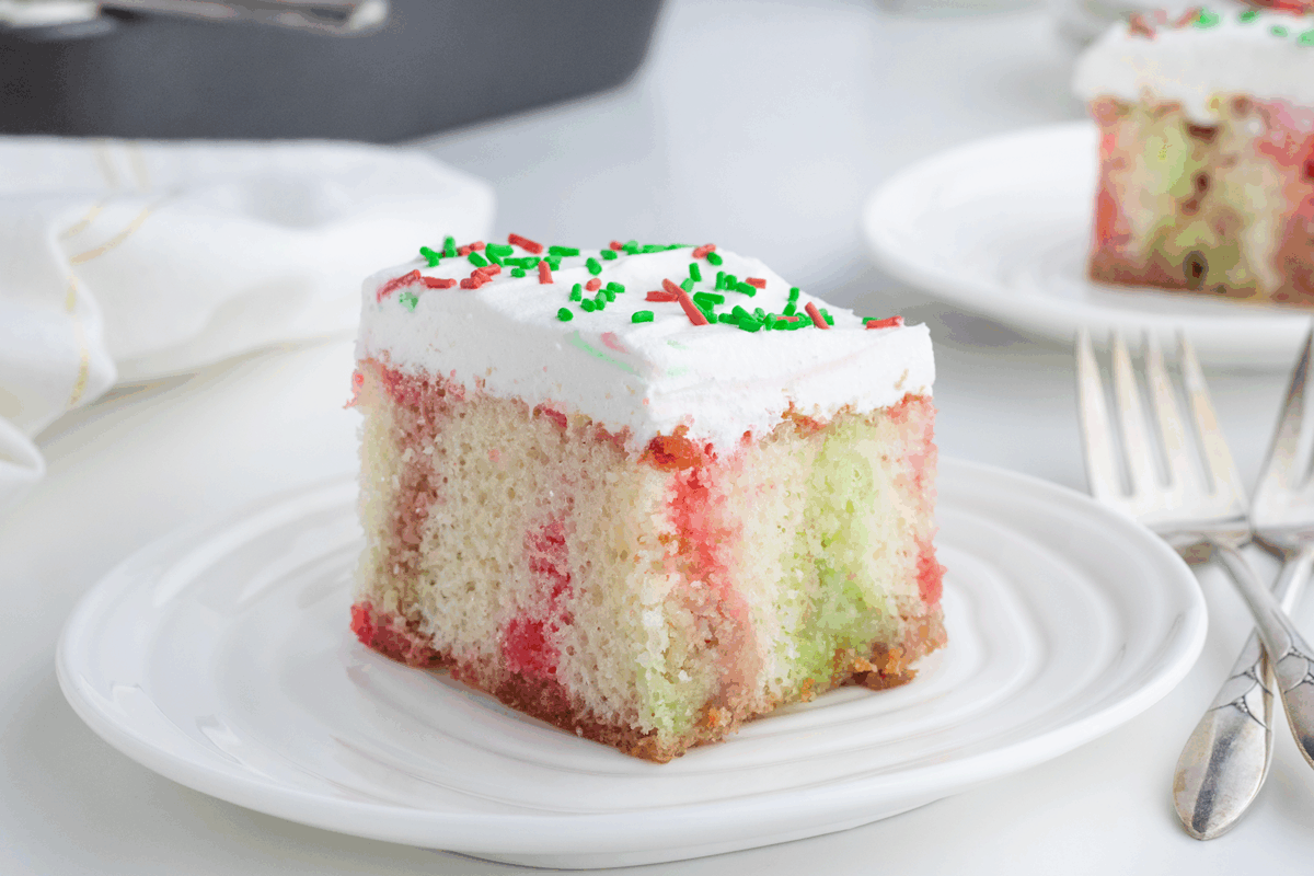 A slice of festive poke cake with red and green streaks, topped with white frosting and red and green sprinkles, sits on a white plate next to a fork and spoon.