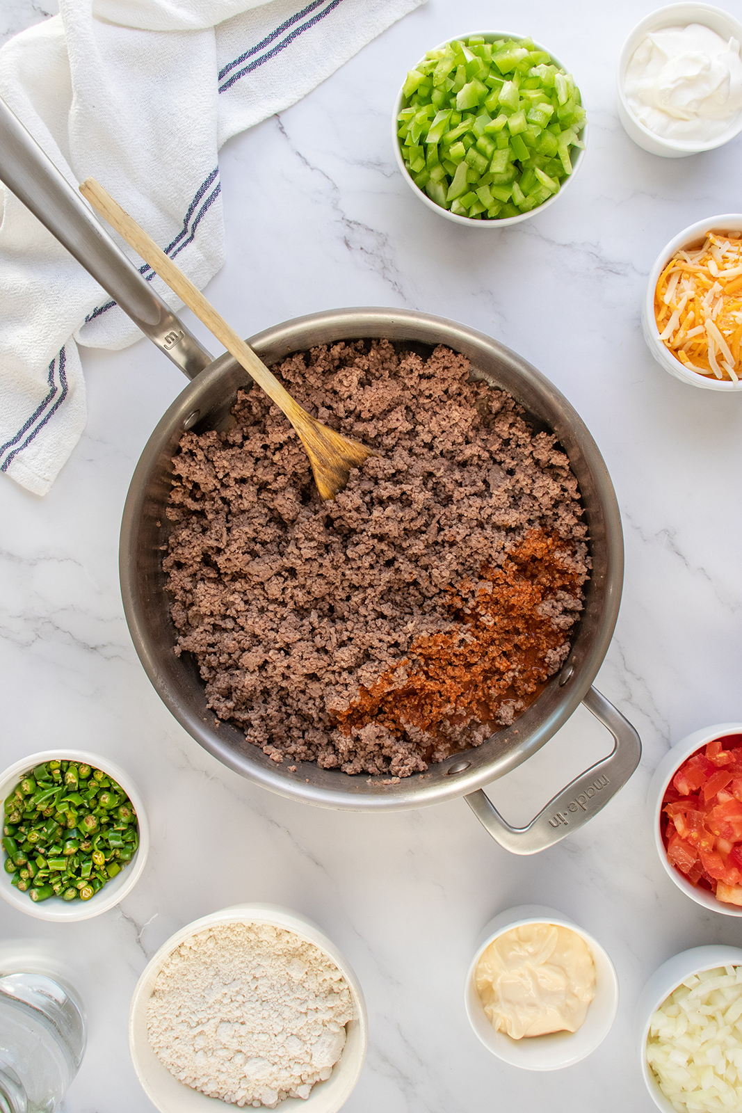 A skillet with browned ground beef and a wooden spoon sits on a marble countertop, surrounded by bowls of chopped green onions, tomatoes, celery, shredded cheese, flour, mayonnaise, and sour cream.