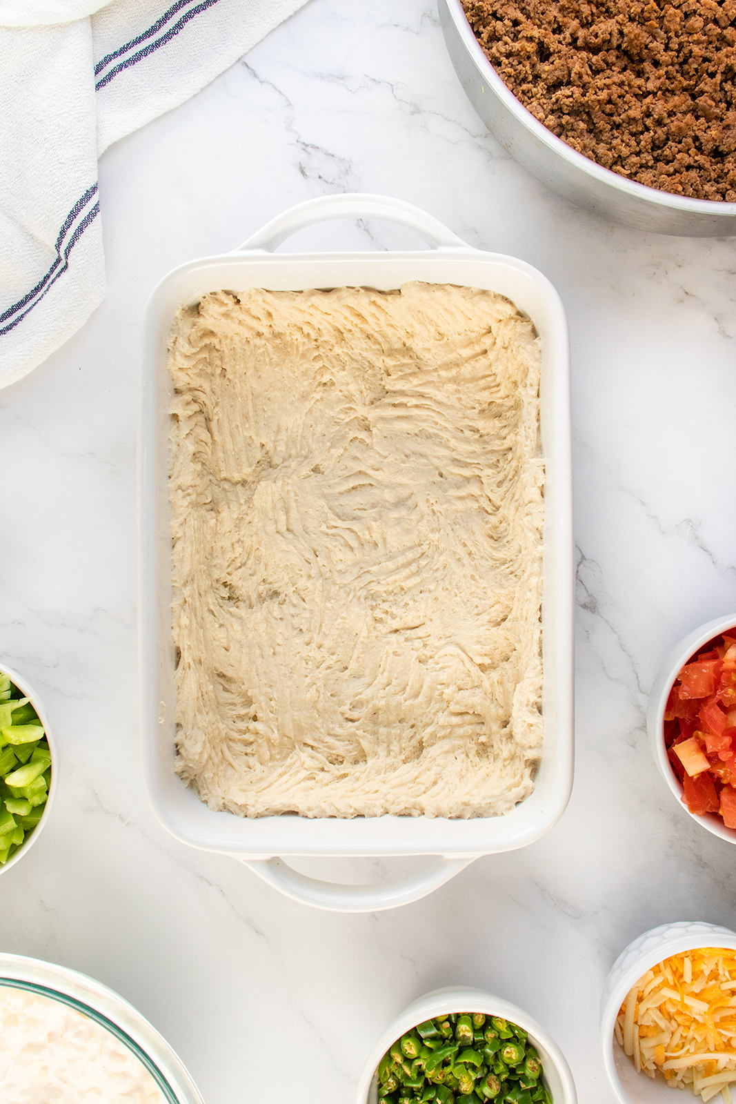 A white baking dish filled with an even layer of mashed potatoes is surrounded by bowls of chopped vegetables, shredded cheese, ground meat, and a striped kitchen towel on a white countertop.