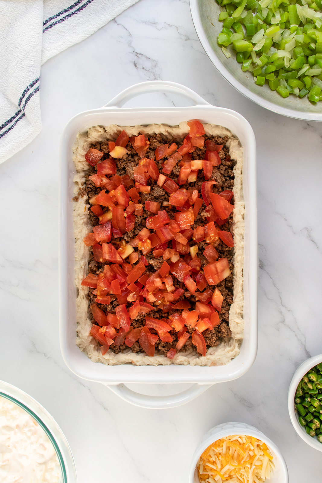 A white baking dish filled with layers of refried beans, cooked ground beef, and chopped tomatoes sits on a marble countertop, surrounded by bowls of shredded cheese, green onions, chopped celery, and a striped towel.