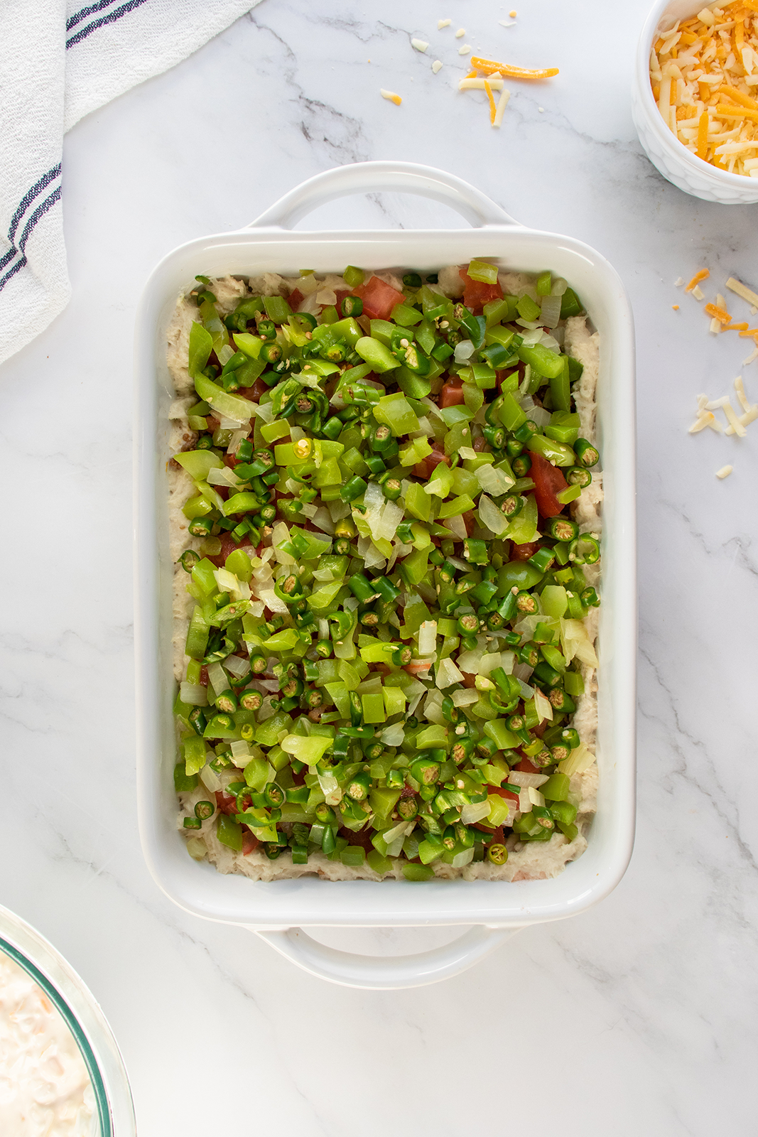 A white rectangular baking dish filled with a mixture of chopped green bell peppers, onions, and tomatoes on a white marble surface. Shredded cheese is visible in a bowl nearby.