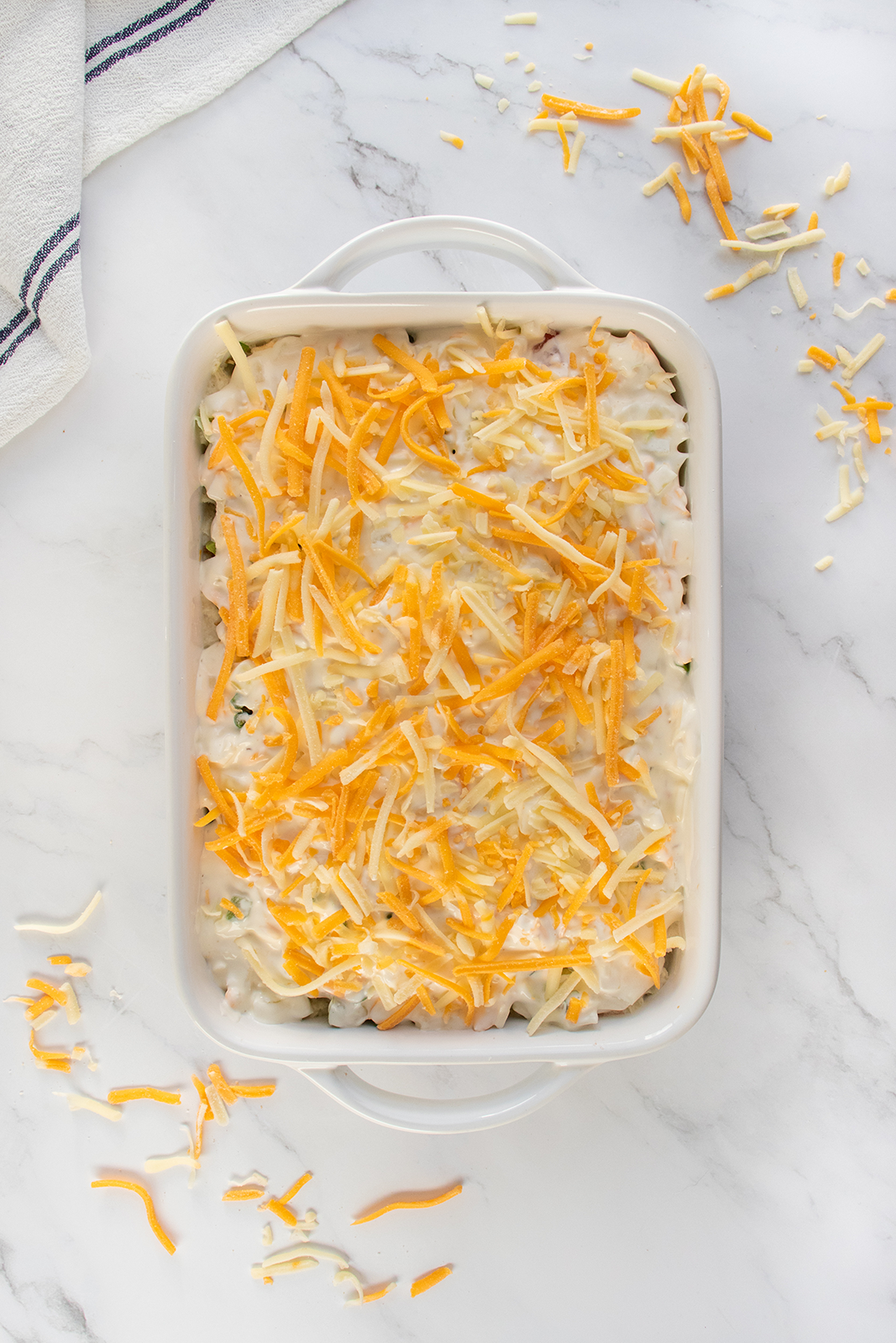 A white baking dish filled with an unbaked casserole topped with shredded yellow and white cheese, sitting on a white marble surface with scattered cheese and a striped kitchen towel nearby.