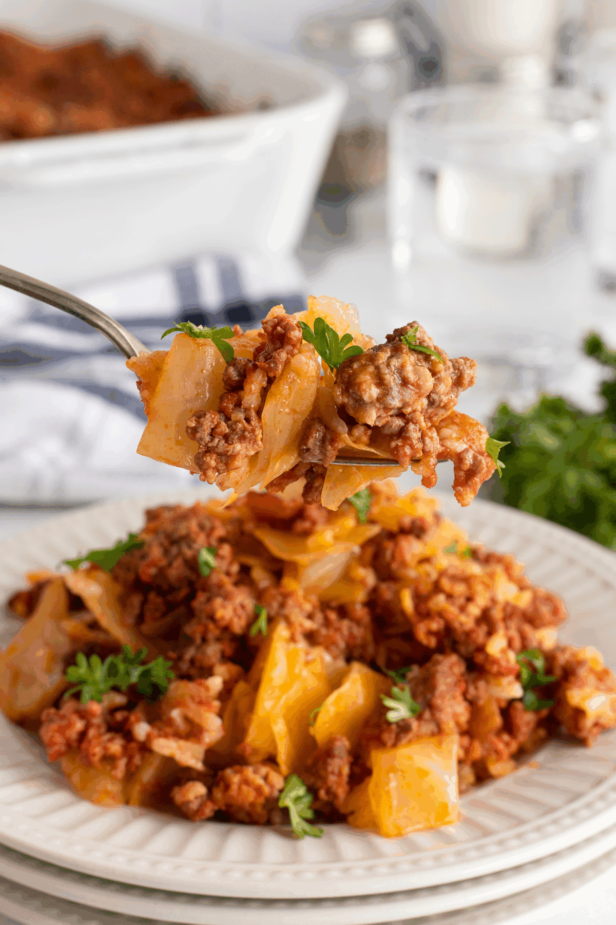 A close-up of a plate of cabbage roll casserole, topped with ground meat and tomato sauce, garnished with parsley. A fork holds a bite above the plate. The background shows a white baking dish and glass of water.