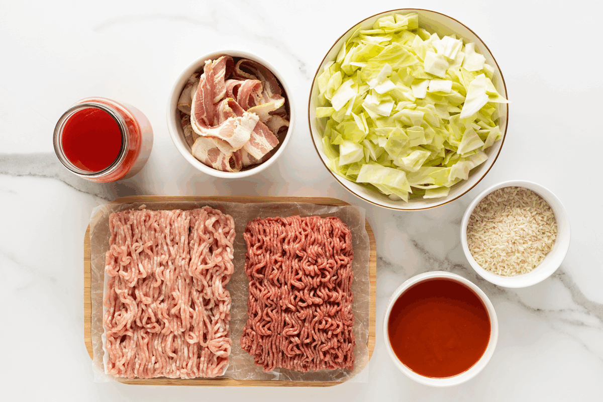 A top-down view of bowls and containers holding raw bacon, chopped cabbage, uncooked rice, tomato sauce, ground pork, and ground beef, all arranged on a white marble surface.