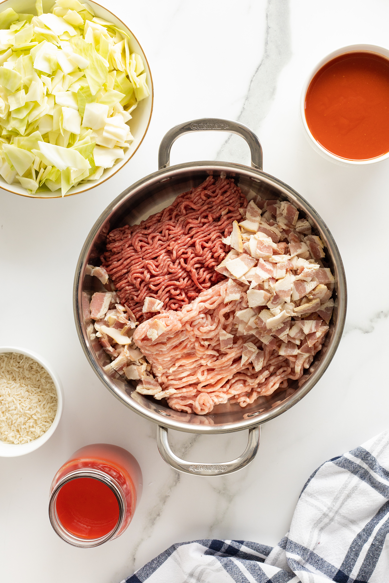 A pot with raw ground beef, ground turkey, and chopped bacon sits on a marble counter, surrounded by bowls of chopped cabbage, rice, tomato sauce, and a jar of tomato sauce, with a striped towel nearby.