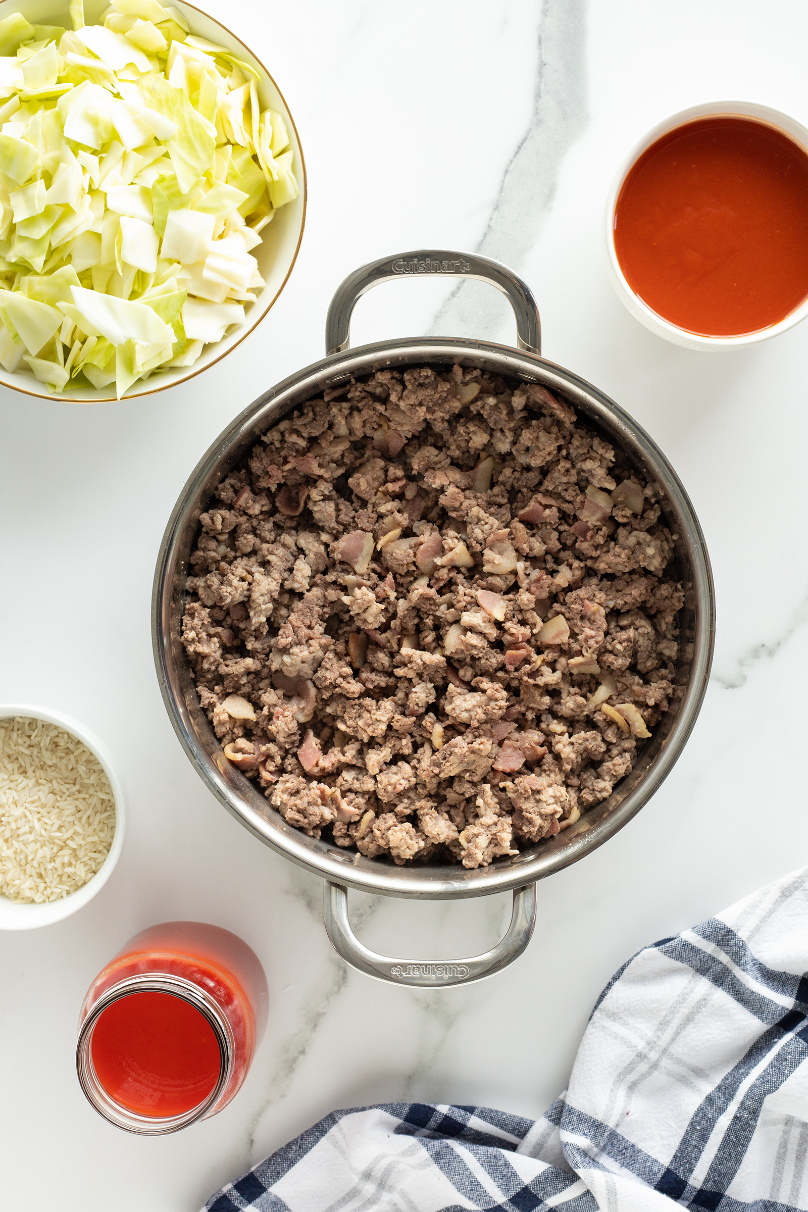 A pot of cooked ground beef sits on a white surface surrounded by bowls containing chopped cabbage, uncooked white rice, and tomato sauce, with a jar of tomato sauce and a striped kitchen towel nearby.
