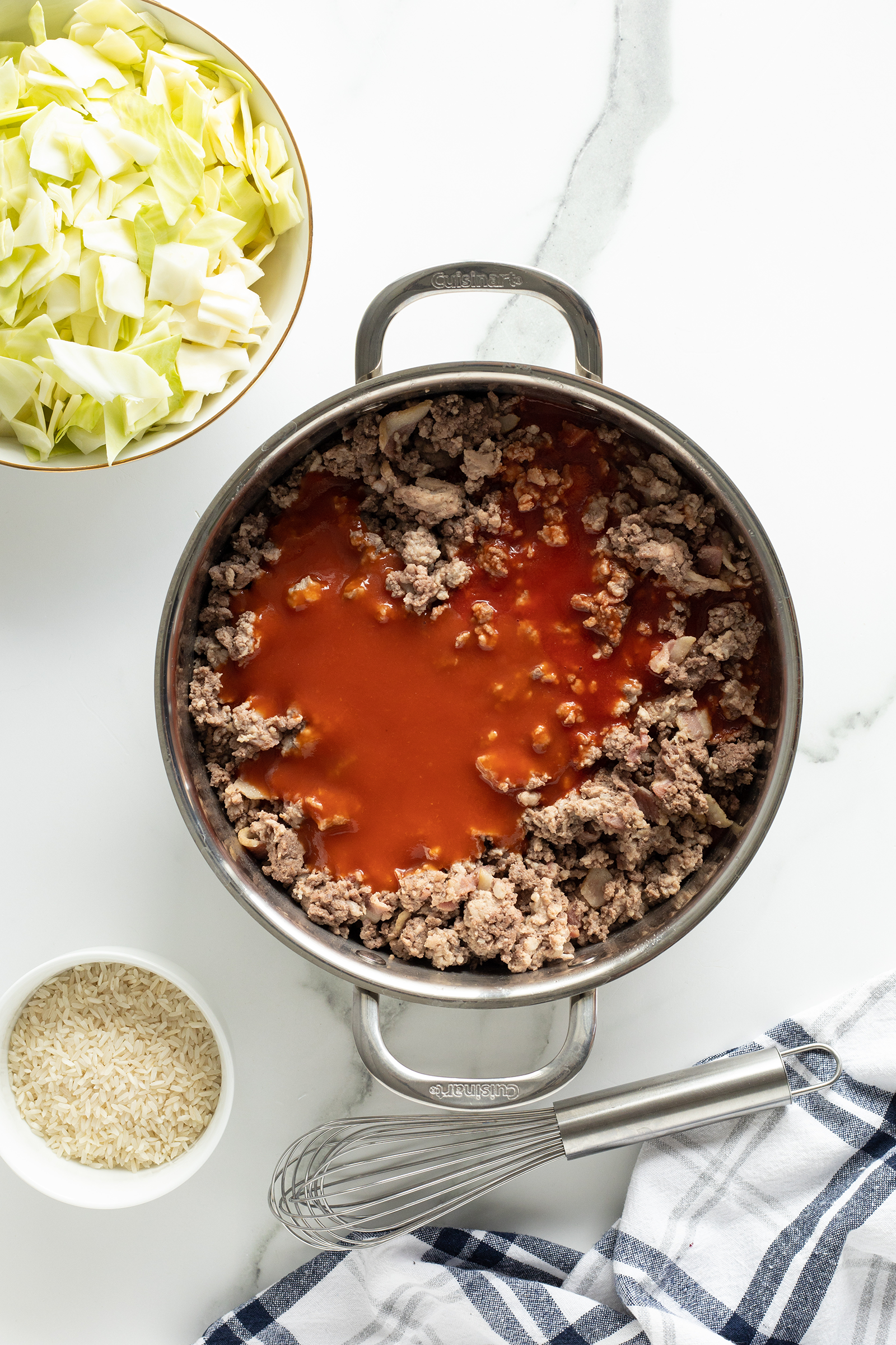 A pot with cooked ground beef and tomato sauce sits on a marble counter, surrounded by a bowl of chopped cabbage, a bowl of uncooked rice, a whisk, and a striped kitchen towel.