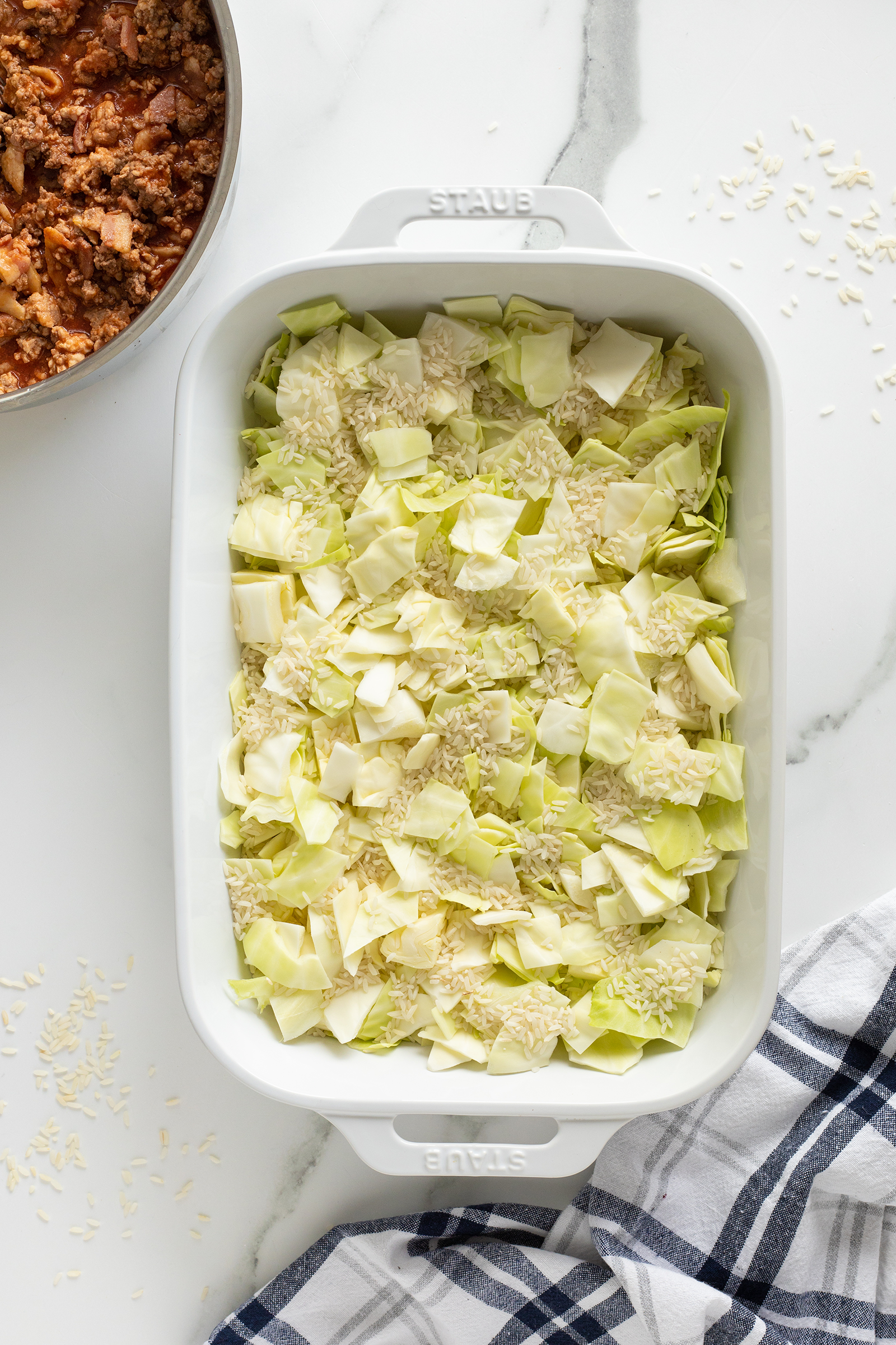 A white baking dish filled with chopped cabbage and uncooked rice sits on a marble countertop, next to a bowl of meat mixture and a blue-and-white striped kitchen towel.